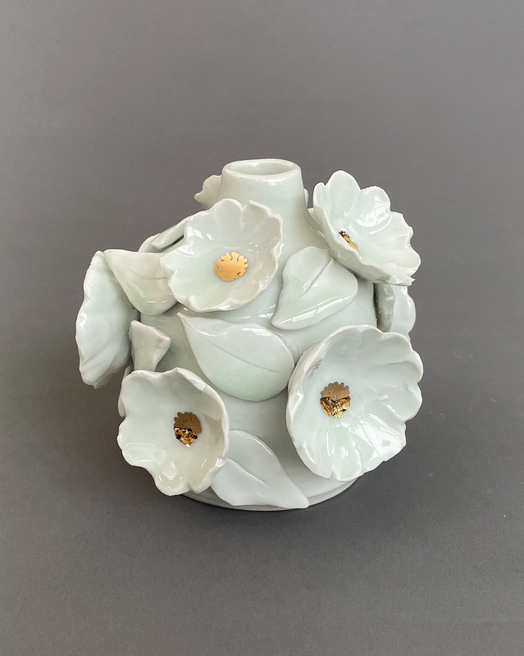 A ceramic vase with white flowers and leaves, decorated with gold accents, on a gray background.