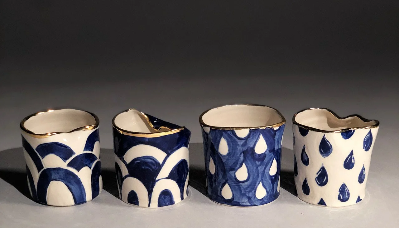 Four small ceramic cups with uneven rims, featuring blue and white patterns, and gold accents along the rims, arranged in a row on a flat surface.