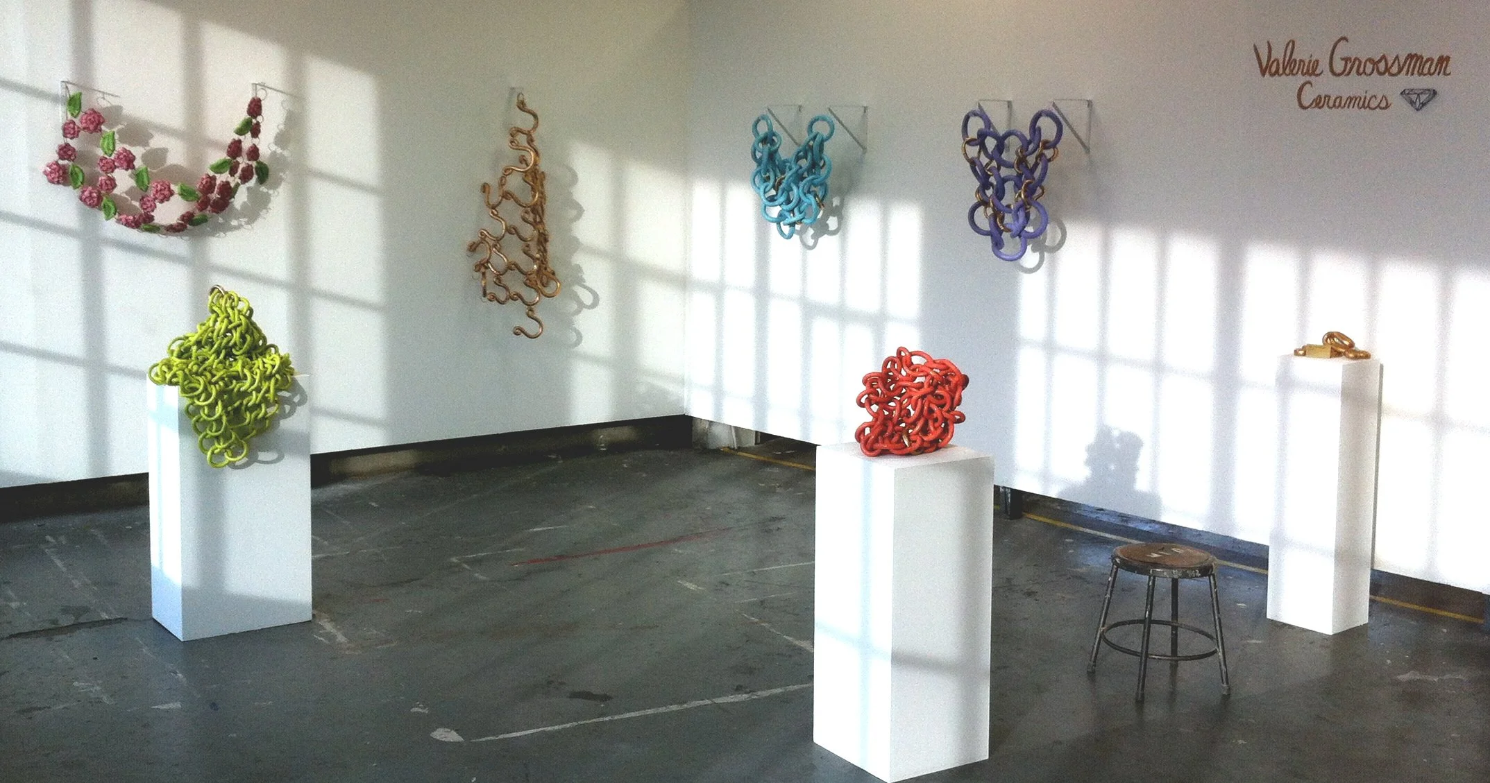 Art exhibit of colorful, abstract sculptures resembling tangled loops and shapes, displayed on white pedestals and wall-mounted panels in a gallery setting.