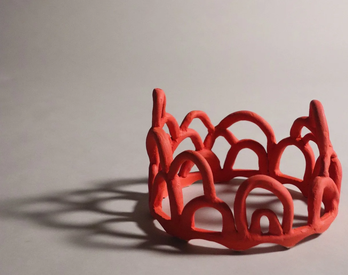 A red, openwork, circular sculpture with arching forms, casting a shadow on a light-colored surface.