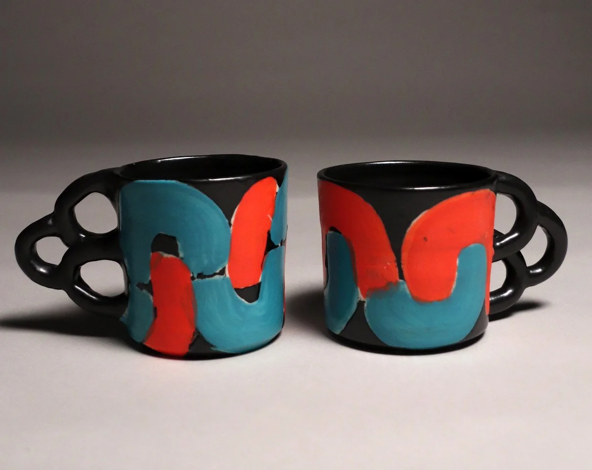 Two black ceramic mugs with colorful abstract red and blue designs and unique looped handles are placed side by side on a light surface against a gray background.