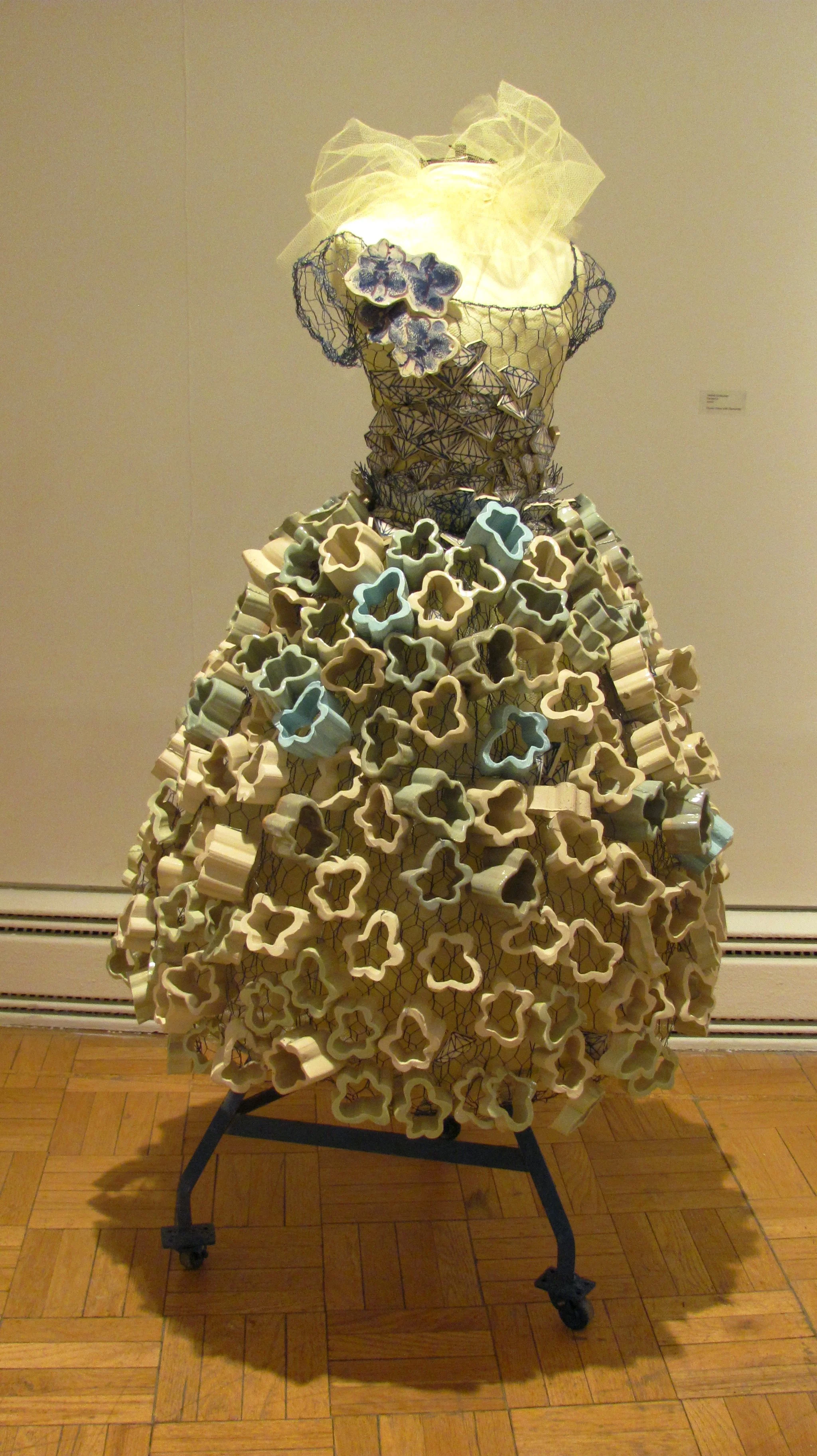 An art installation of a dress made from multiple extruded ceramic flowers with cutouts, arranged in a layered fashion, with a lit頭 and a sheer fabric on top, displayed in an art gallery.