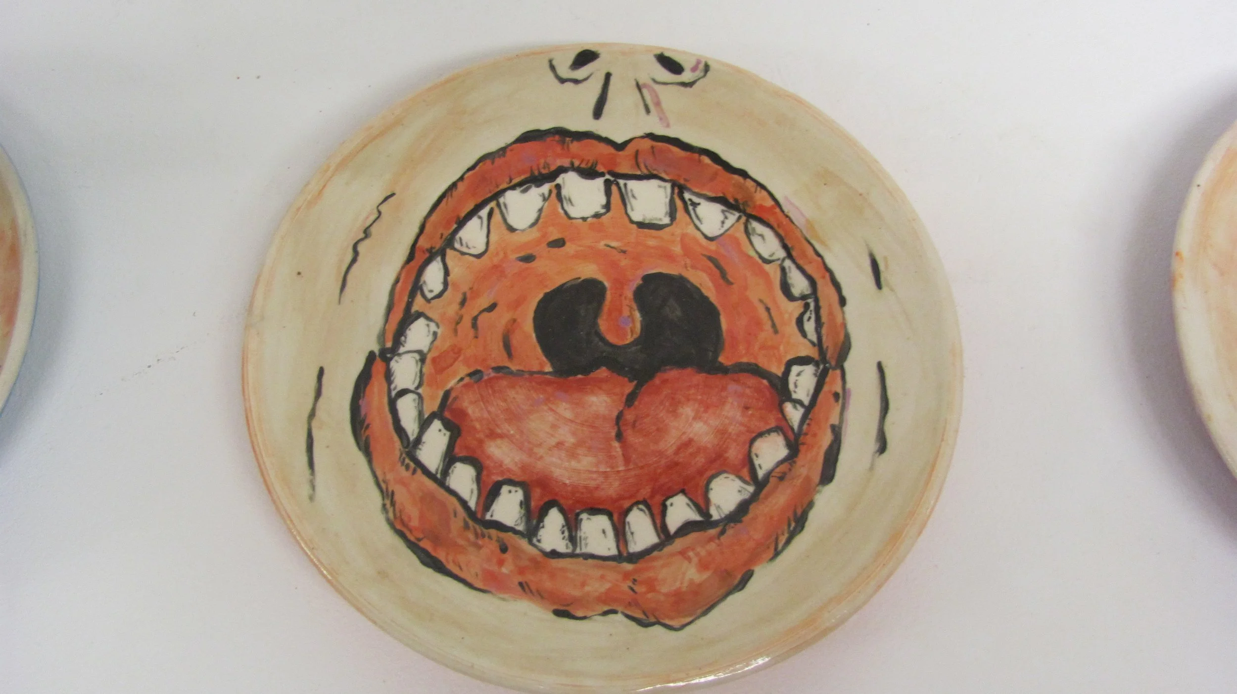 A ceramic plate with a painted drawing of an open mouth with teeth and a tongue, illustrating a cartoonish face.