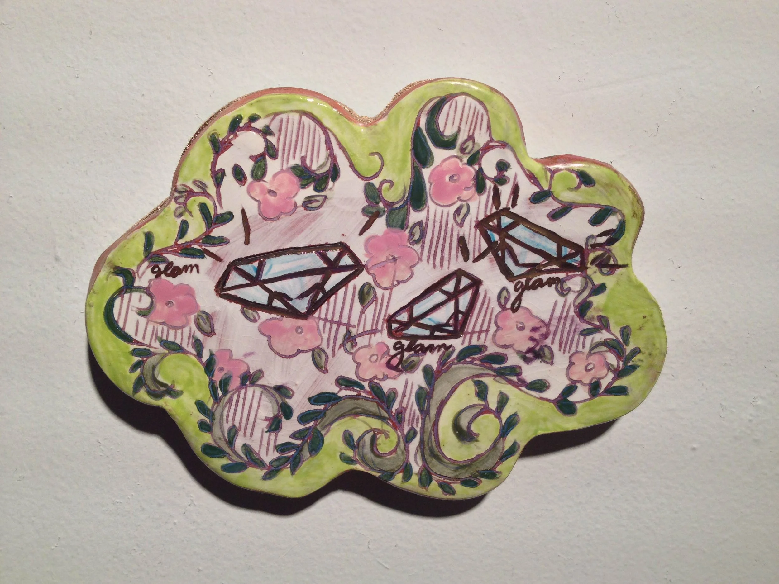 Decorative ceramic plate with pink flowers, green leaves, and illustrations of diamonds labeled 'glam' on a light background.