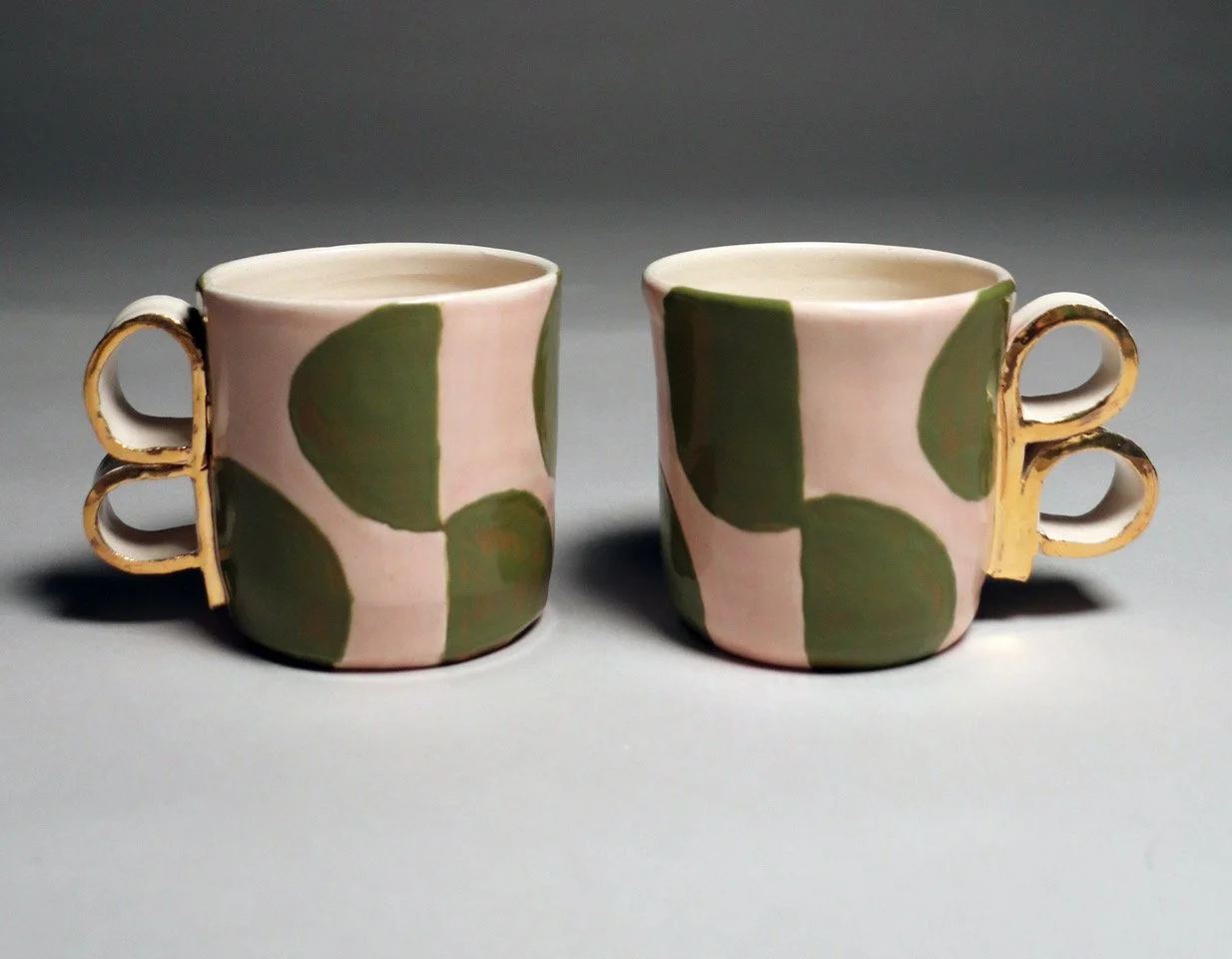 Two ceramic mugs with pink and green floral patterns and gold handles placed side by side on a gray surface.