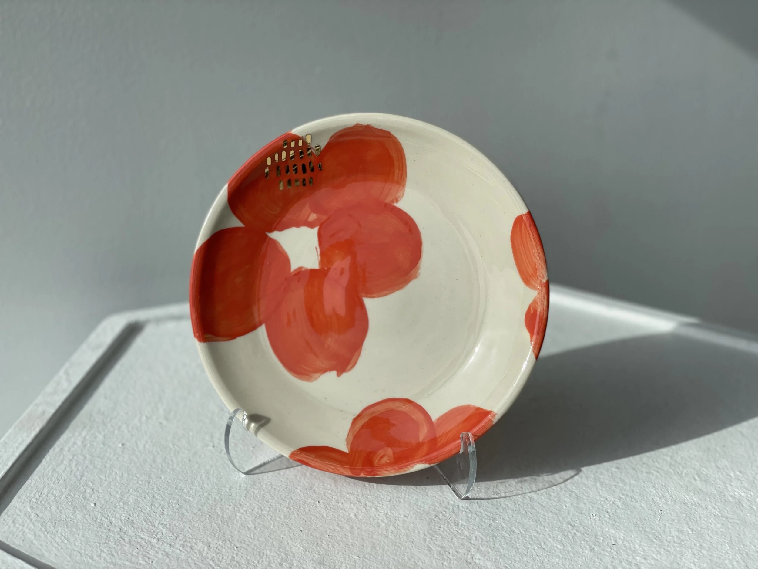 Ceramic bowl with a white background and red abstract floral pattern, displayed on a clear plastic stand on a white surface, with a light gray wall background.