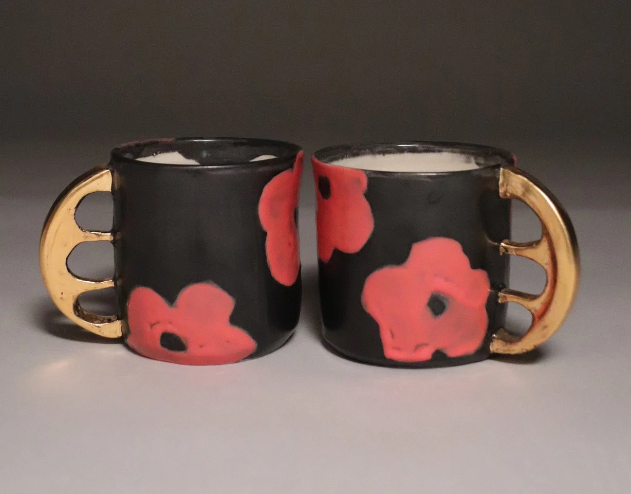 Two black cups with gold handles and pink floral patterns, placed side by side on a gray surface with a gray background.