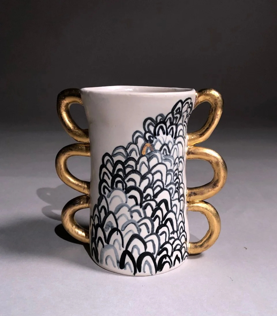 A small ceramic mug with black and gold wave-like patterns and gold-colored handles on each side.