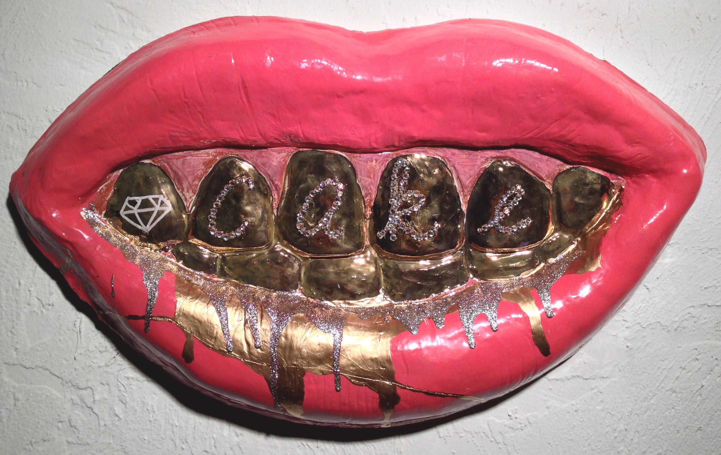 A large wall art piece shaped like lips with gold teeth spelling out the word "cake" in diamond-like letters, with pink, gold, and glitter accents on a white textured wall.