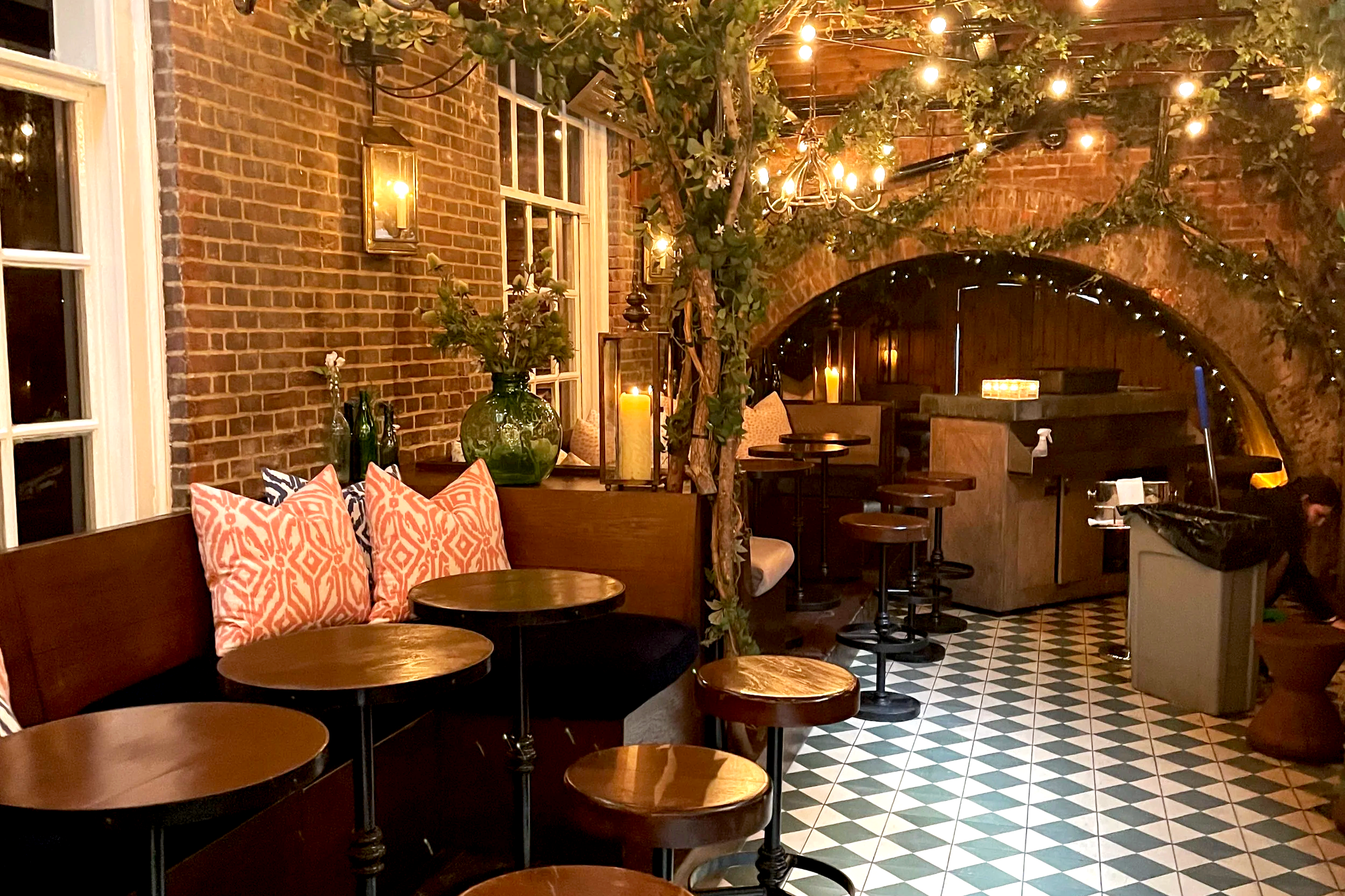 Cozy restaurant interior with exposed brick walls, wooden furniture, patterned tile flooring, decorative pillows, green vases with flowers, and string lights hanging from ceiling with greenery