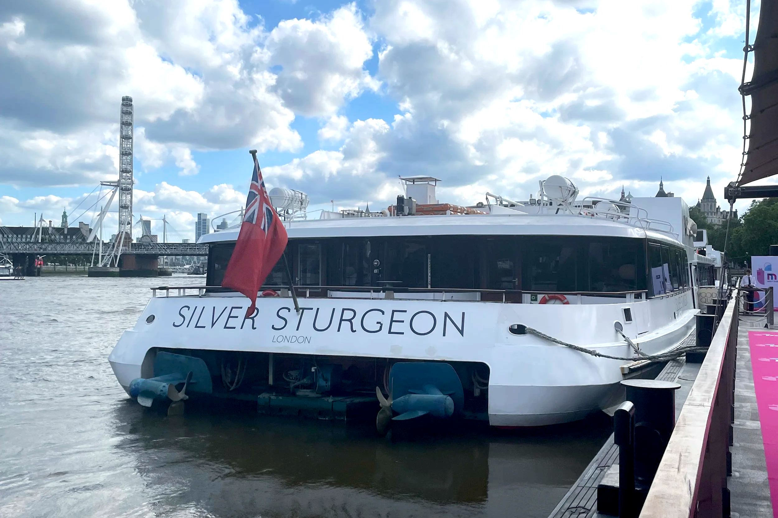 A white passenger boat named 'Silver Sturgeon' docked along the riverbank with London skyline and clouds in the background. The boat has a Union Jack flag on the front.