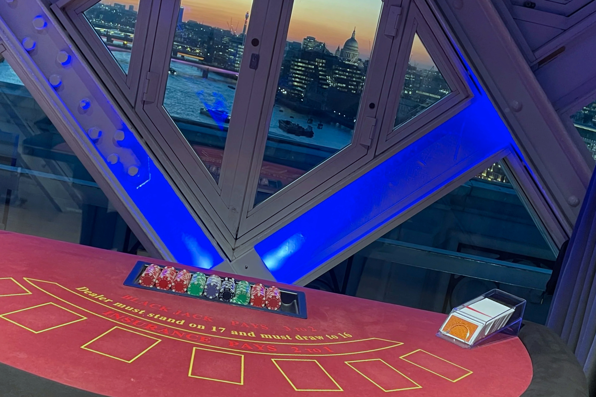 Inside a casino with a roulette table, colorful chips, and a view of the city skyline including St. Paul's Cathedral at sunset through large windows with blue accent lighting.