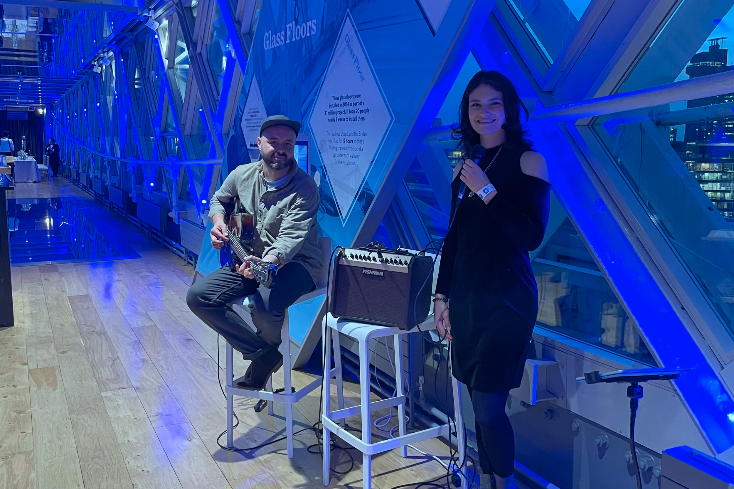 A man playing an acoustic guitar and a woman holding a microphone stand in a modern, blue-lit indoor space with glass floors and structural design elements, possibly a public venue or event space.