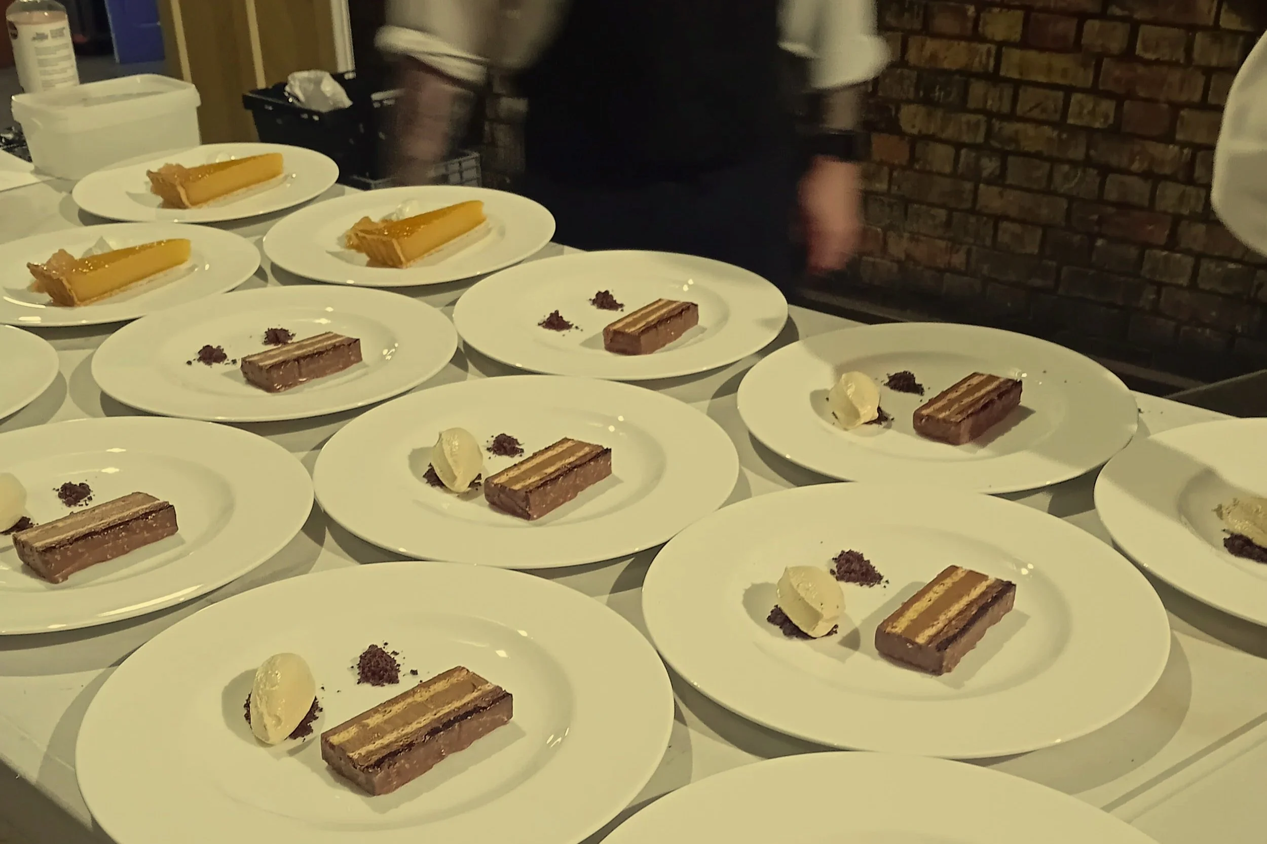 Multiple white plates with slices of layered cakes, chocolate with cream, and white ice cream, arranged on a table, with a person in the background and a brick wall.