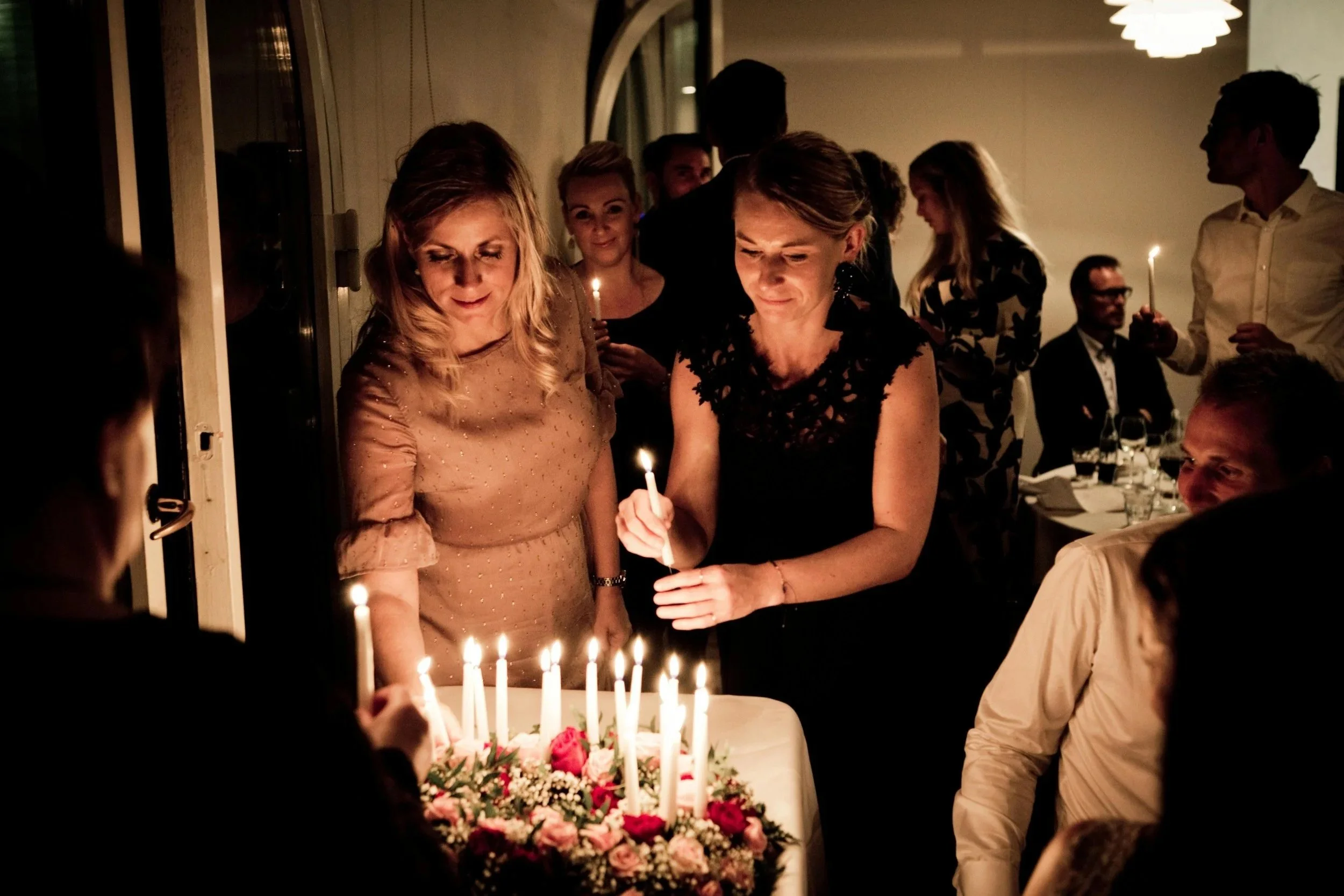 Group of people celebrating a birthday with a cake decorated with flowers and lit candles, some holding sparklers at night.