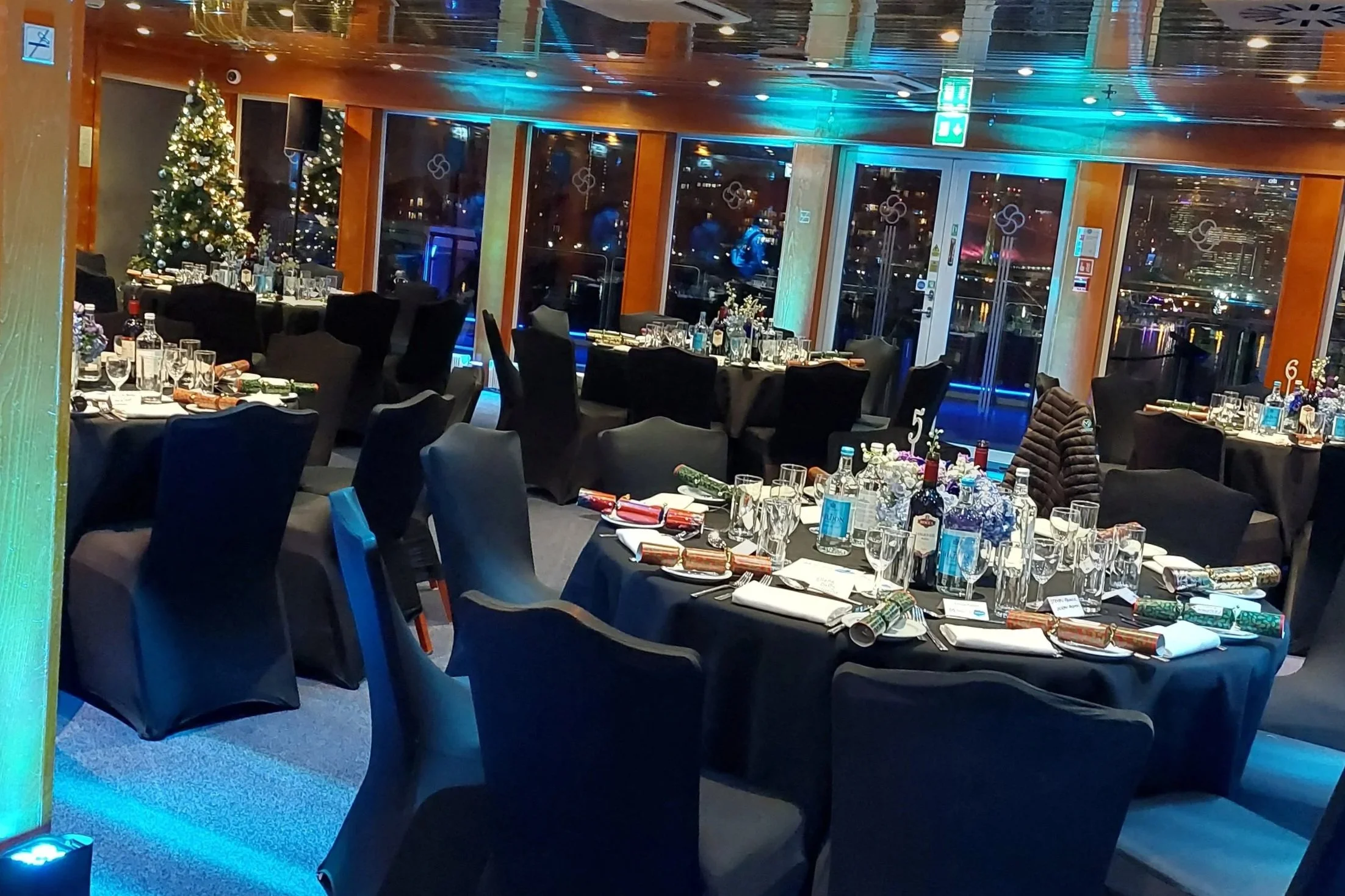 Elegant dining room decorated for Christmas with black tablecloths, tall Christmas trees with lights, and neatly arranged glassware and silverware on the tables, overlooking a cityscape at night through large windows.
