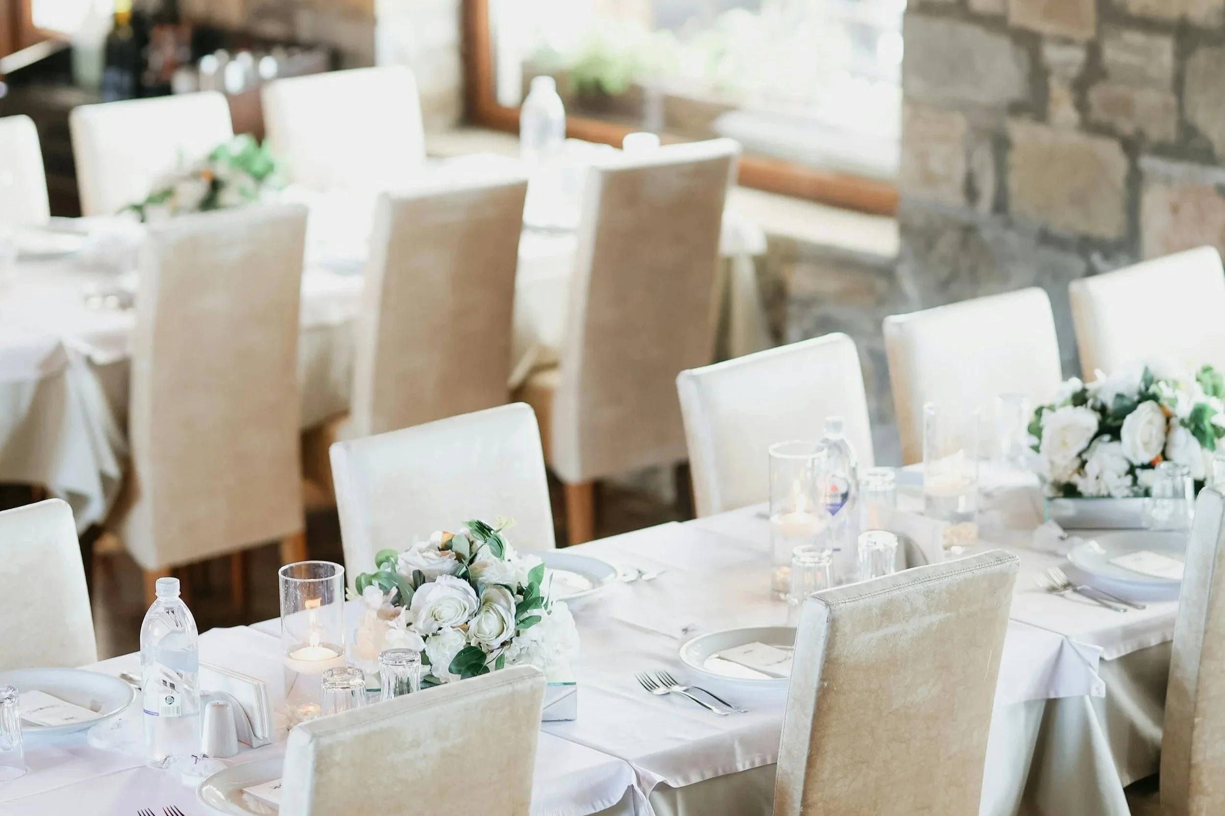 A dining area set for a special occasion with white tablecloths, floral centerpieces, candles, water bottles, and neatly arranged plates and silverware in a bright room with large windows.