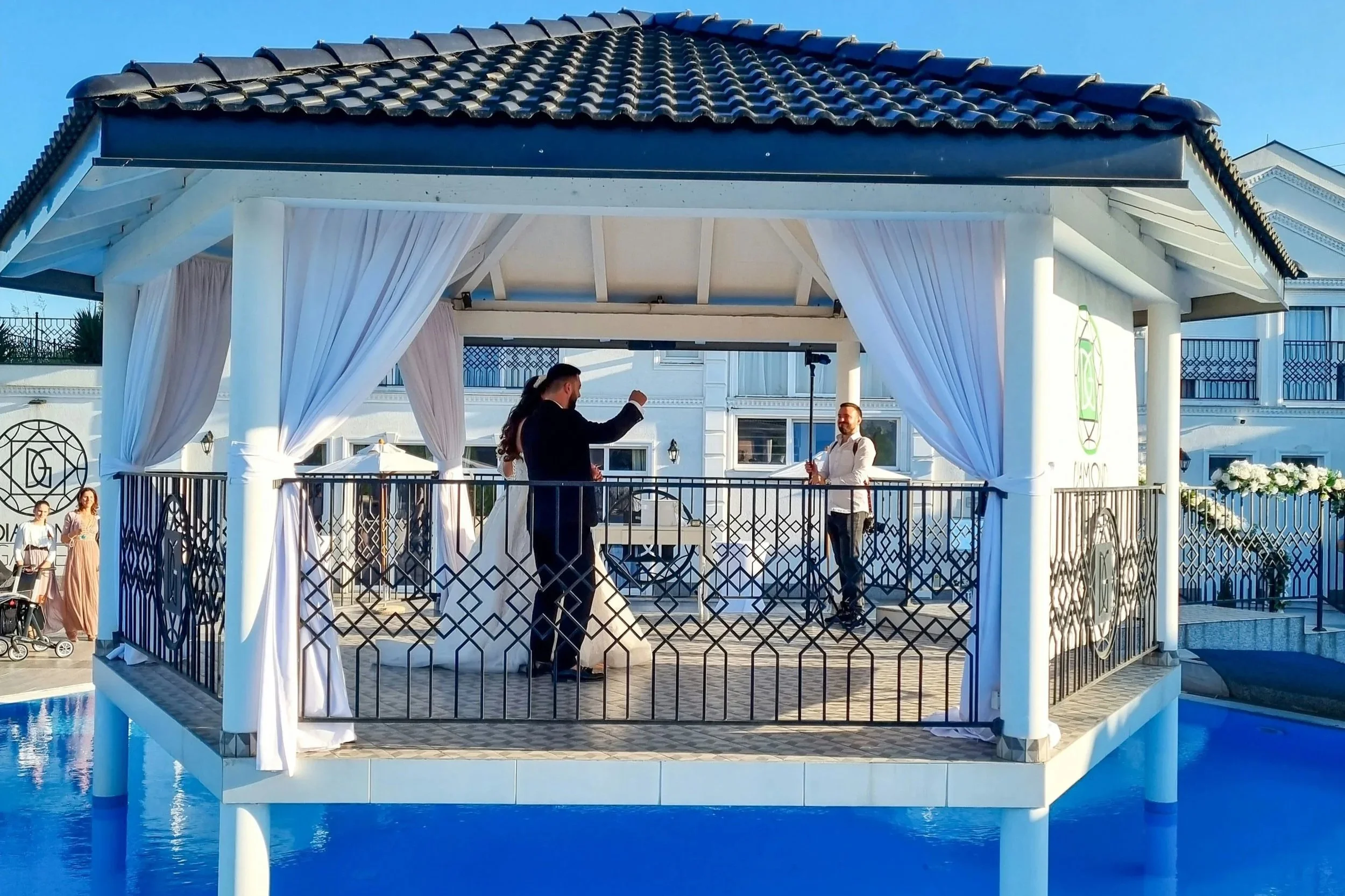 A couple getting married on a gazebo built over a swimming pool, with a man and a woman dressed in wedding attire, surrounded by white curtains. There are two women with strollers and children in the background.