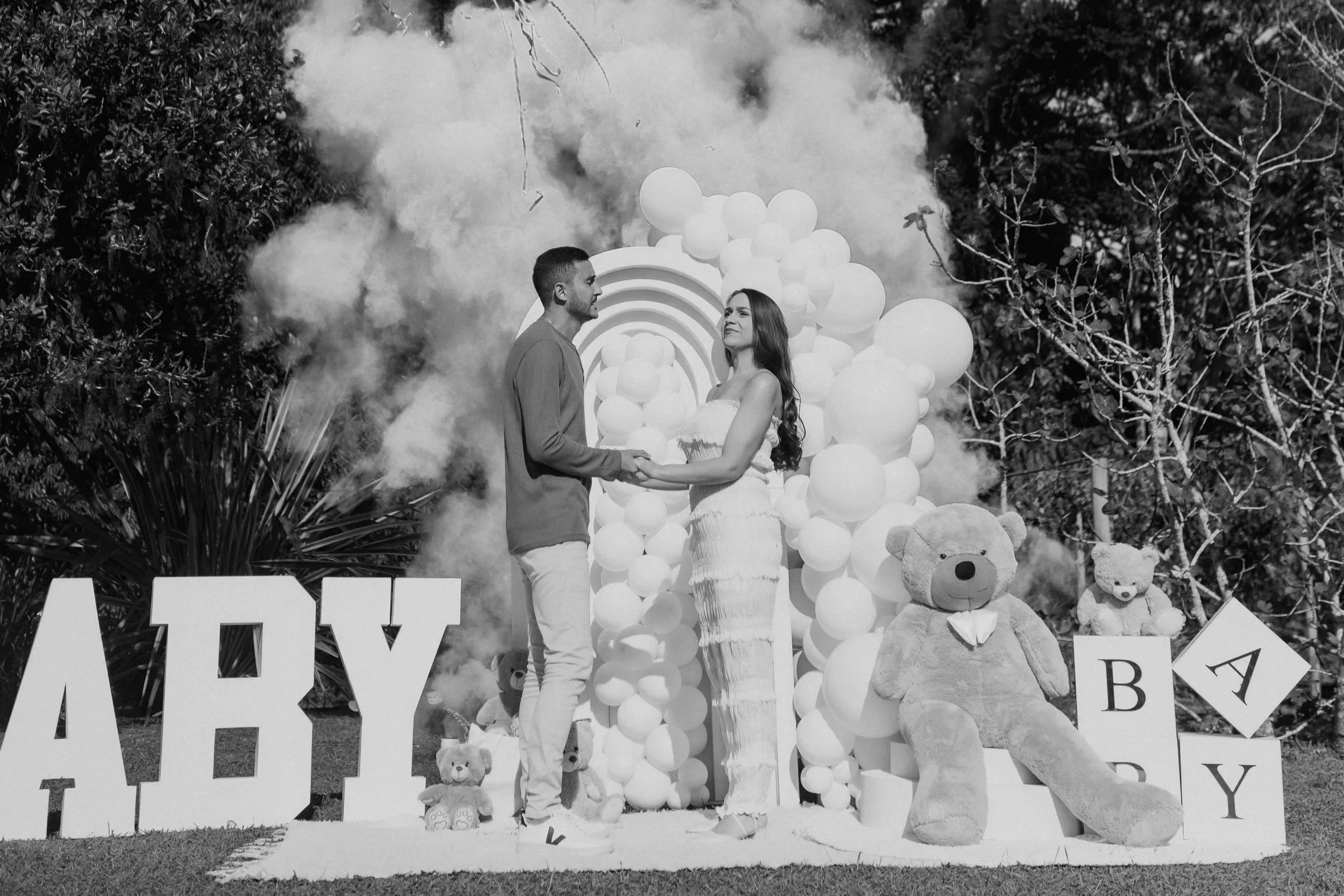 A couple stands holding hands during a baby shower outdoors, with a balloon arch, large teddy bears, and blocks spelling 'BABY' in the background. Smoke or fog adds a decorative atmosphere.