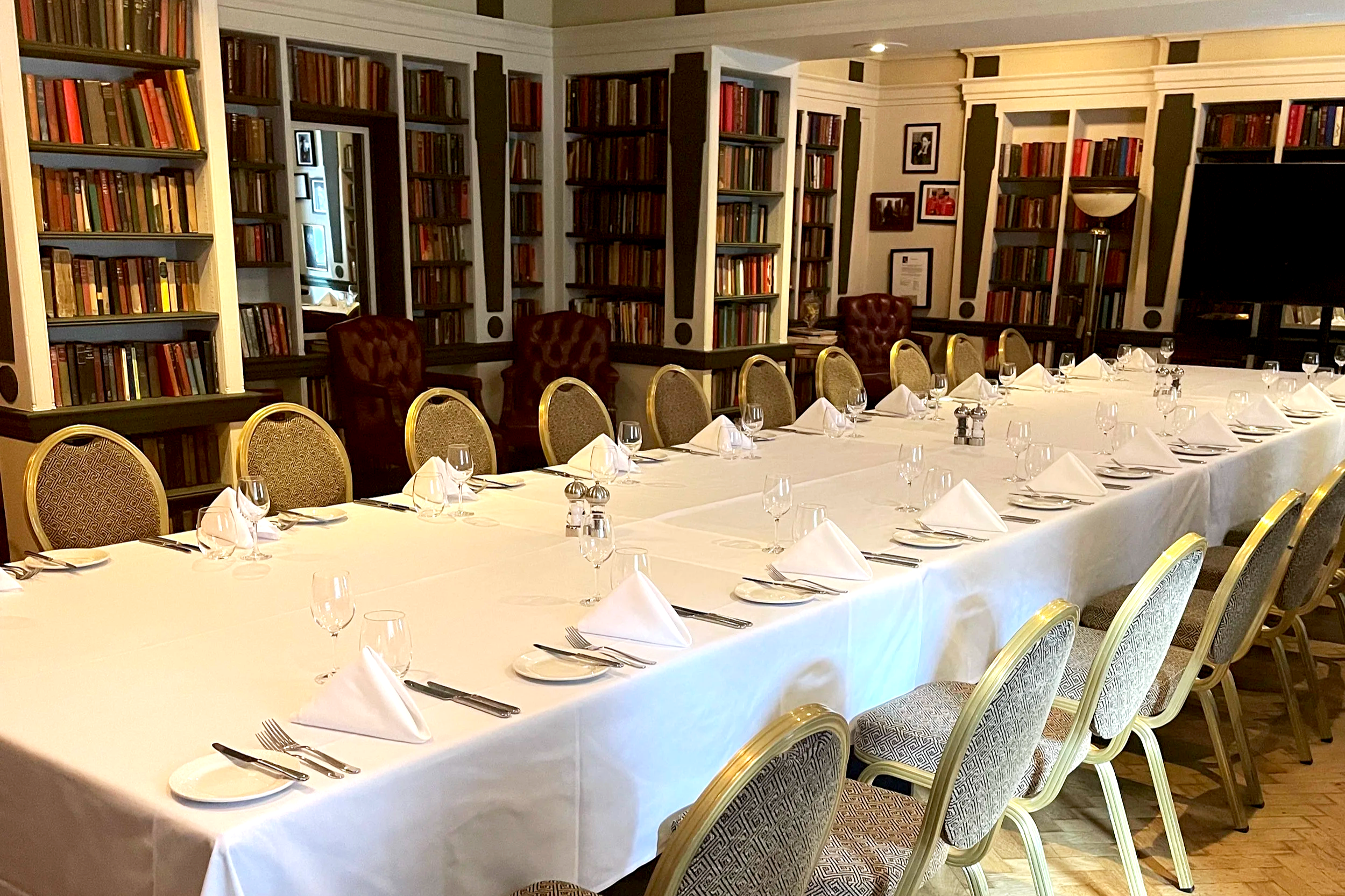 A long dining table set for a formal meal in a library-themed room with bookshelves, framed photographs, and a large TV.