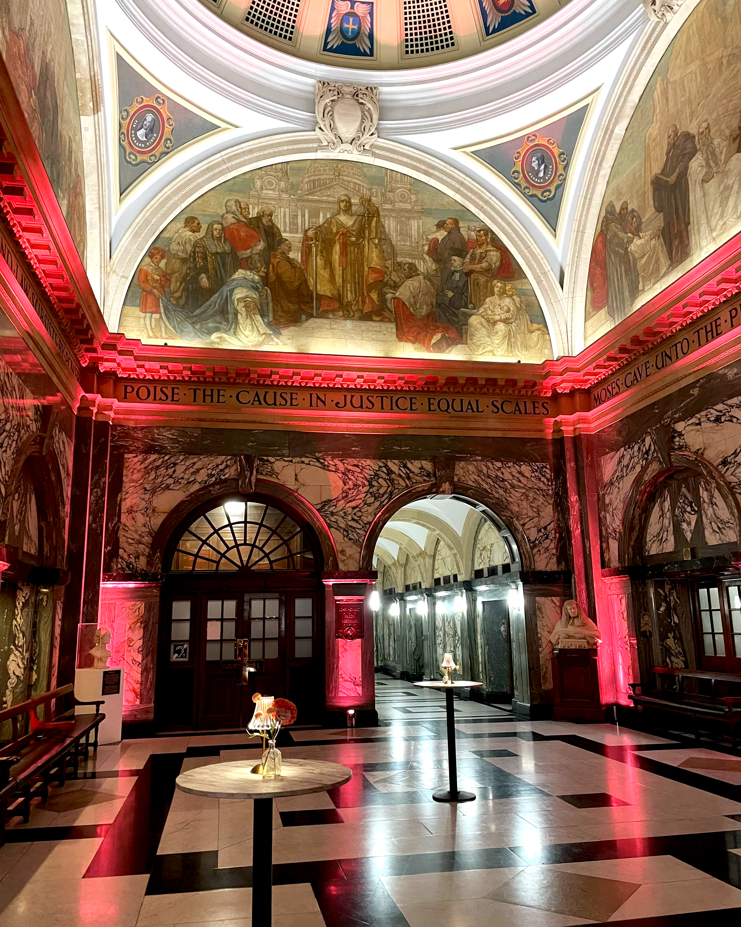 Interior of a grand historic building with marble floors, pink lighting, and artwork on the walls. There are small tables with lamps and a mural of a historical event on the ceiling.