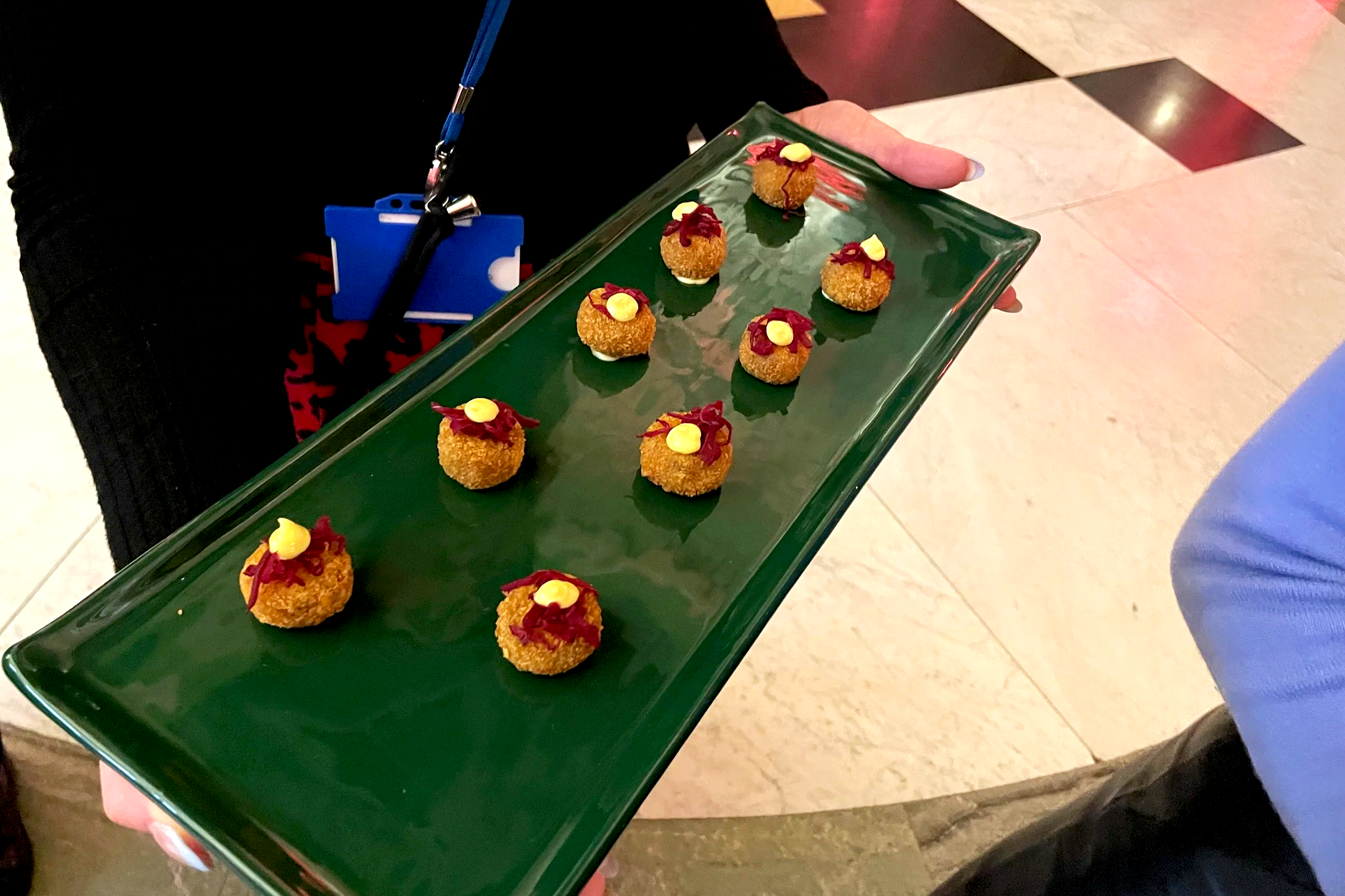 A person holding a green rectangular tray with ten small, round, breaded appetizers topped with red sauce and small yellow garnish, in an indoor setting with a tiled floor.