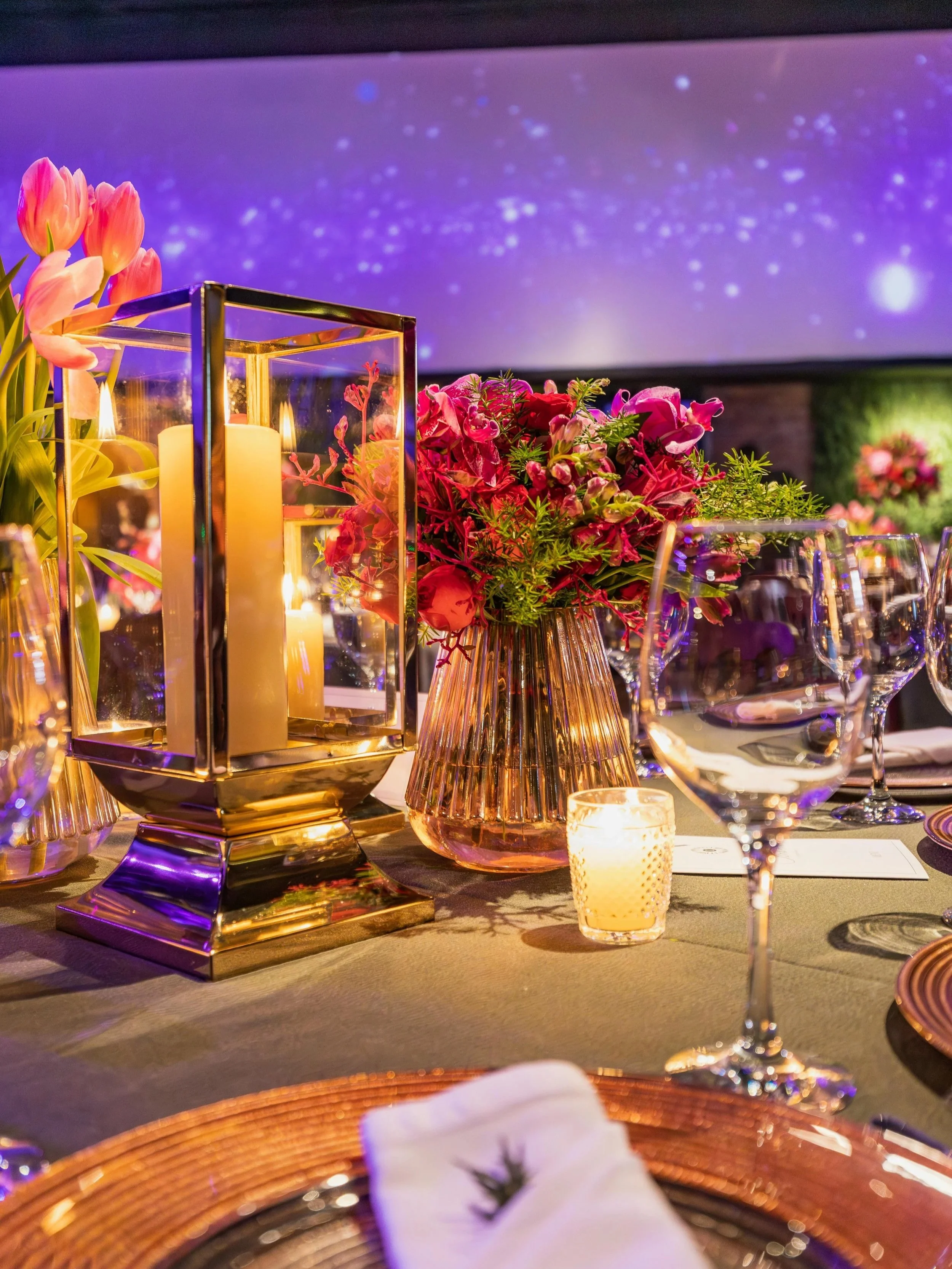 Elegant table centerpiece with pink flowers, a gold lantern with candles, and a small votive candle, set for a formal event with wine glasses and table settings under purple lighting.