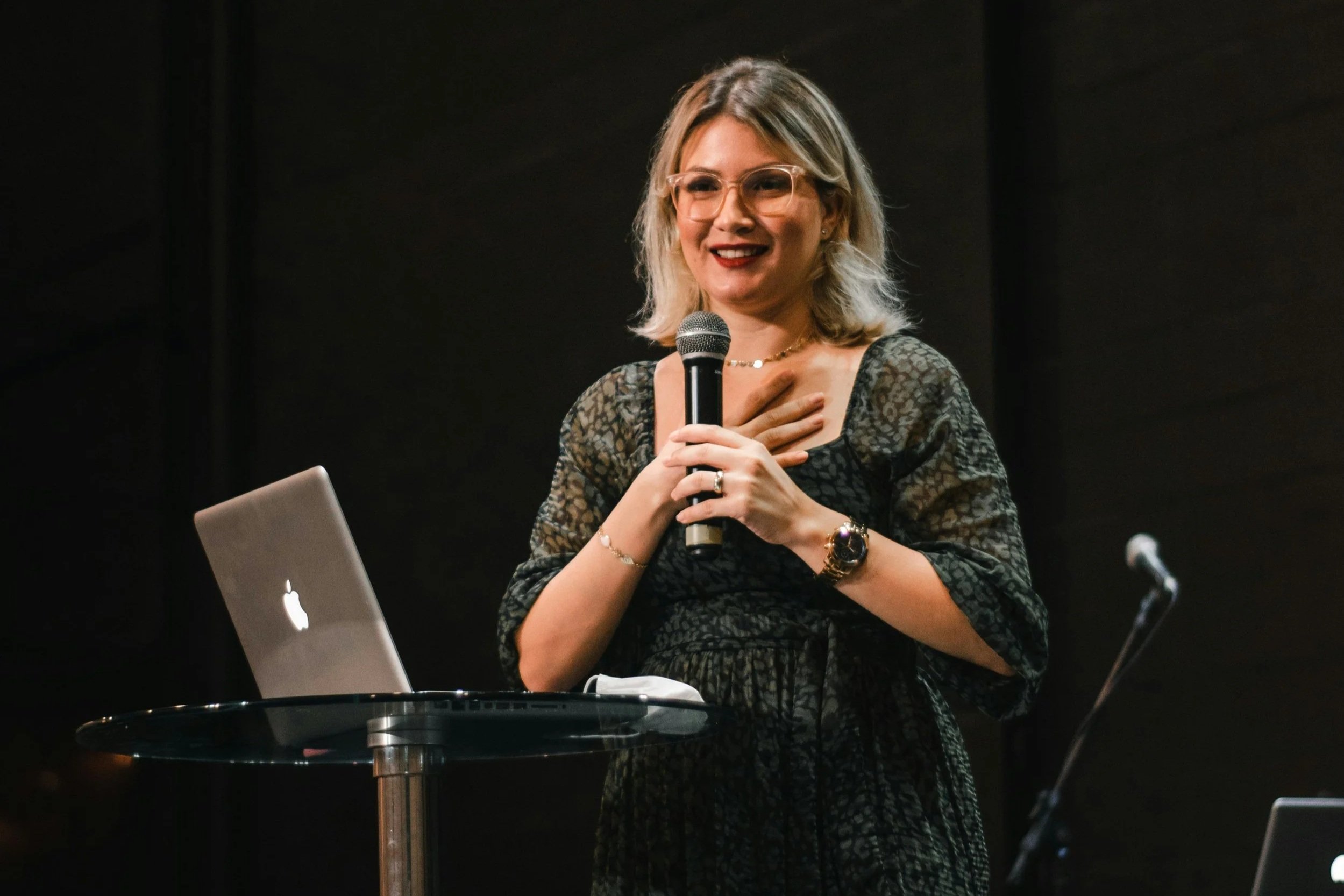 A woman with blonde hair wearing glasses, a black dress with lace pattern, holding a microphone and standing behind a small round table with a silver MacBook laptop. She has her left hand over her heart and is smiling.