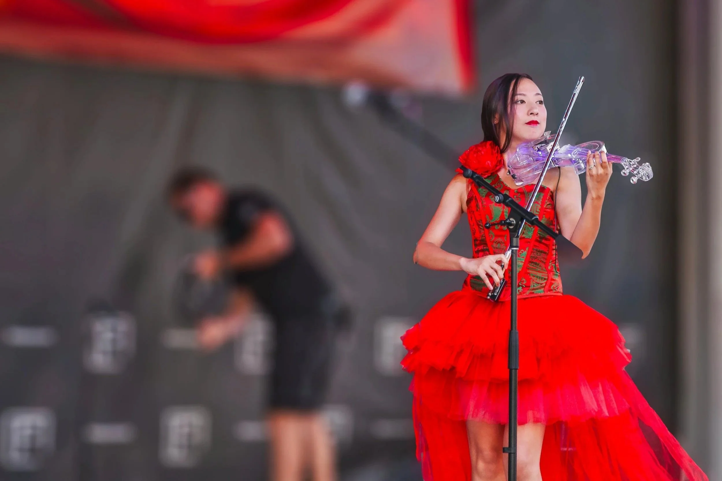 A woman in a red dress playing a violin on stage, with a blurred person in the background adjusting equipment.
