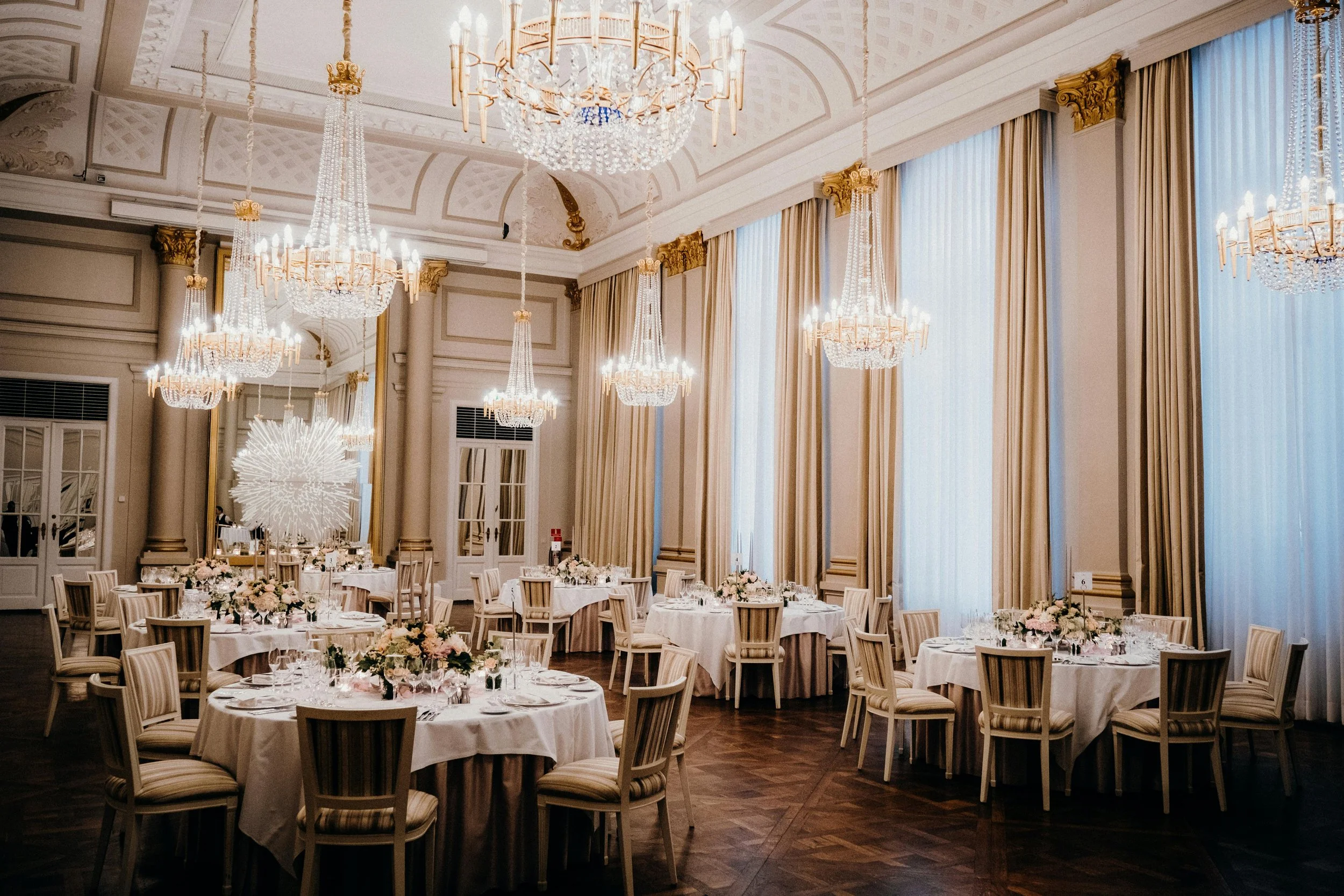 Elegant banquet hall with large chandeliers, tall draped curtains, round tables with white tablecloths, floral centerpieces, and wooden chairs, ready for an event.
