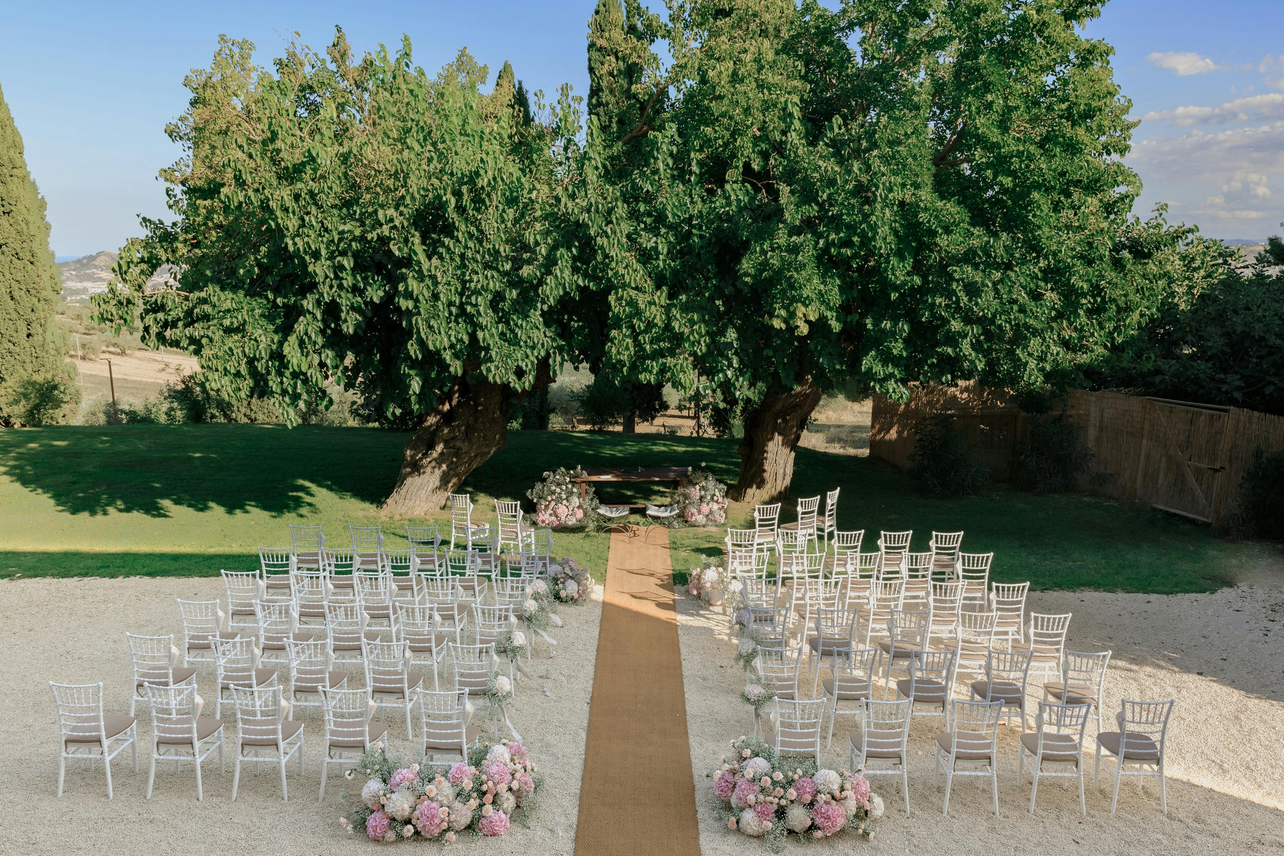Outdoor wedding ceremony setup with white chairs arranged in rows on a grass and gravel surface, decorated with pink and white flowers, leading to an altar under two large trees with lush green foliage, in a scenic natural setting.