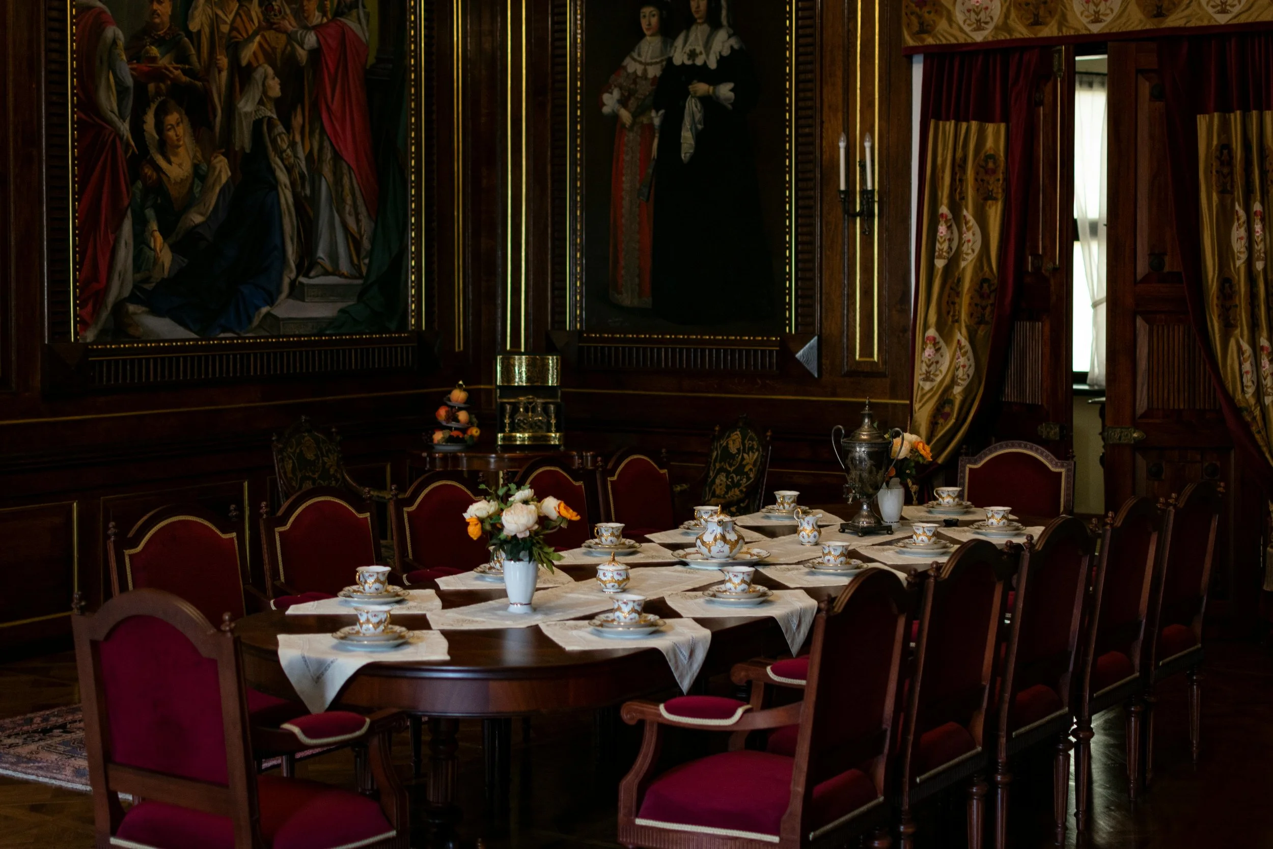 Elegant dining room with a long wooden table set with fine china, teacups, and saucers, decorated with a vase of flowers, surrounded by red velvet chairs, with ornate paintings on the dark wood-paneled wall and draped curtains.