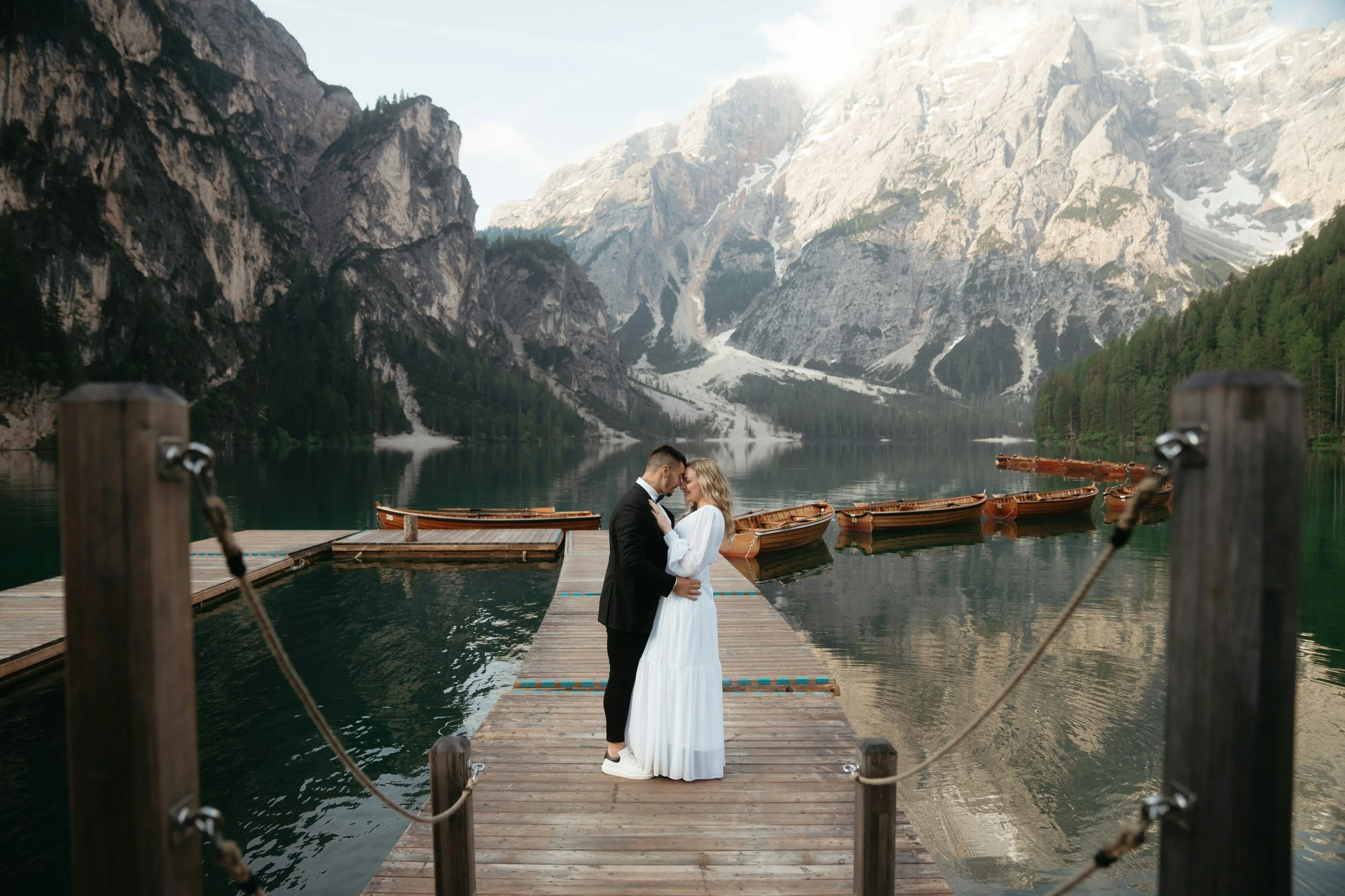 A couple embracing on a wooden dock by a lake surrounded by mountains and forests, with boats moored nearby.