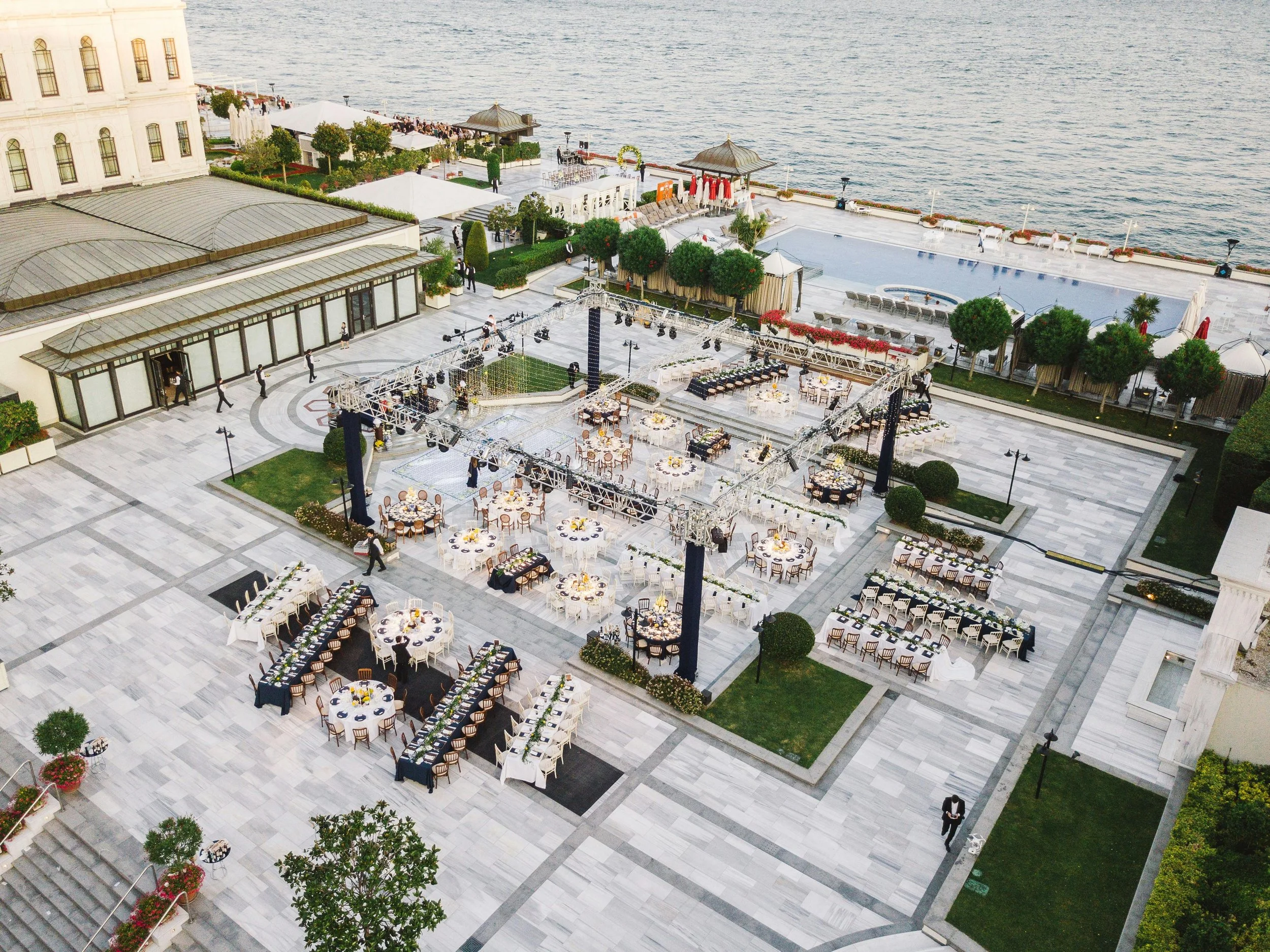 Aerial view of an outdoor event setup with decorated tables, lighting, and a stage near a waterfront.