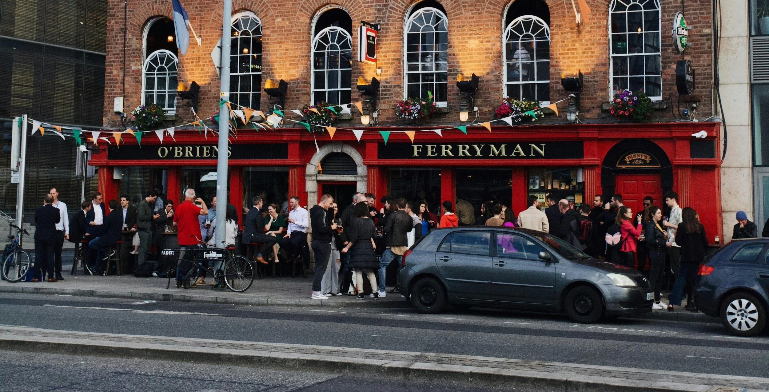 Crowd of people gathered outside a pub named O'Brien's Ferryman on a busy city street, some standing and talking, others sitting at tables, with cars parked along the curb and colorful pennant banners decorating the building.
