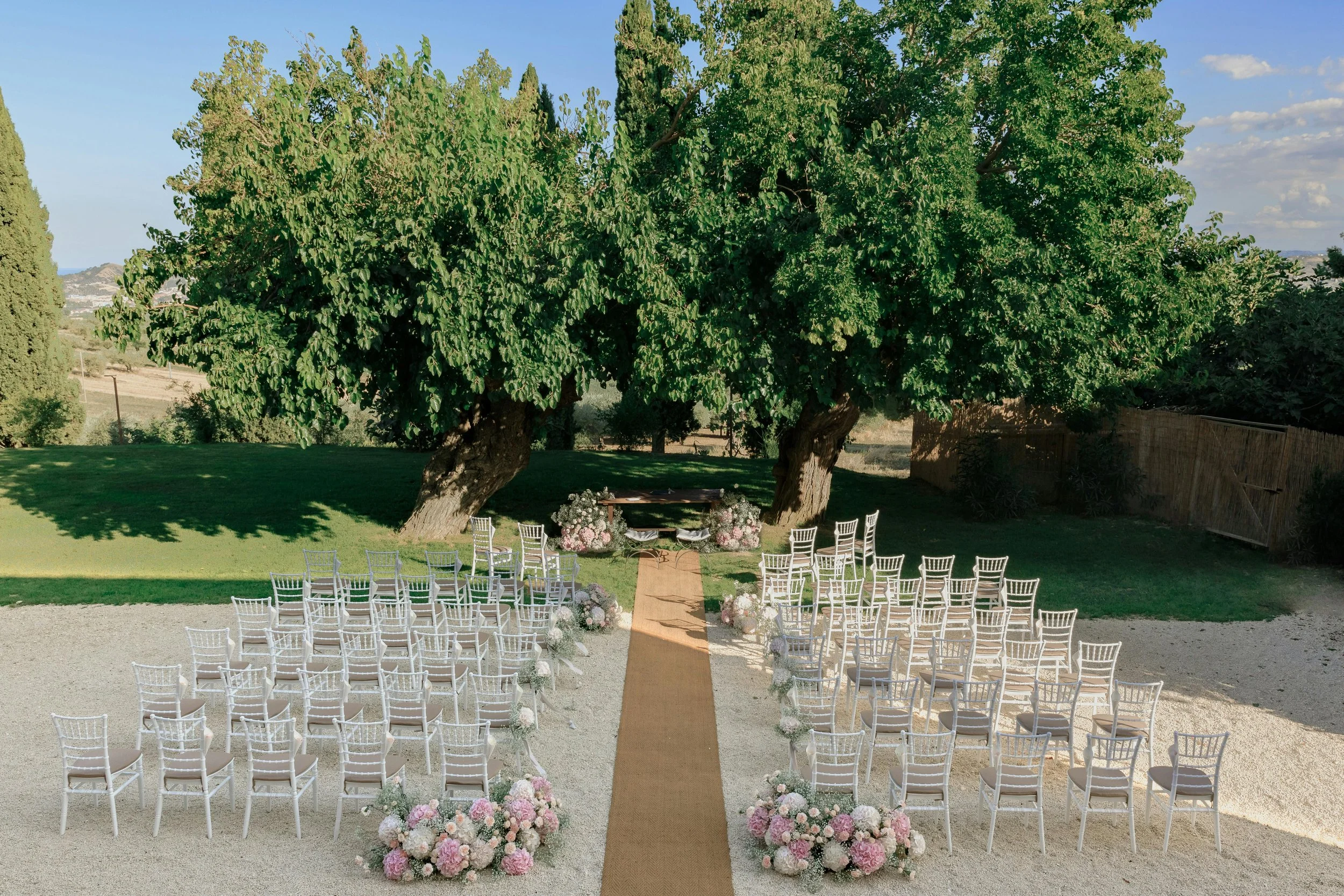 Outdoor wedding ceremony setup under large trees with white chairs arranged in rows, an aisle decorated with floral arrangements, and a table at the front with more flowers on either side.