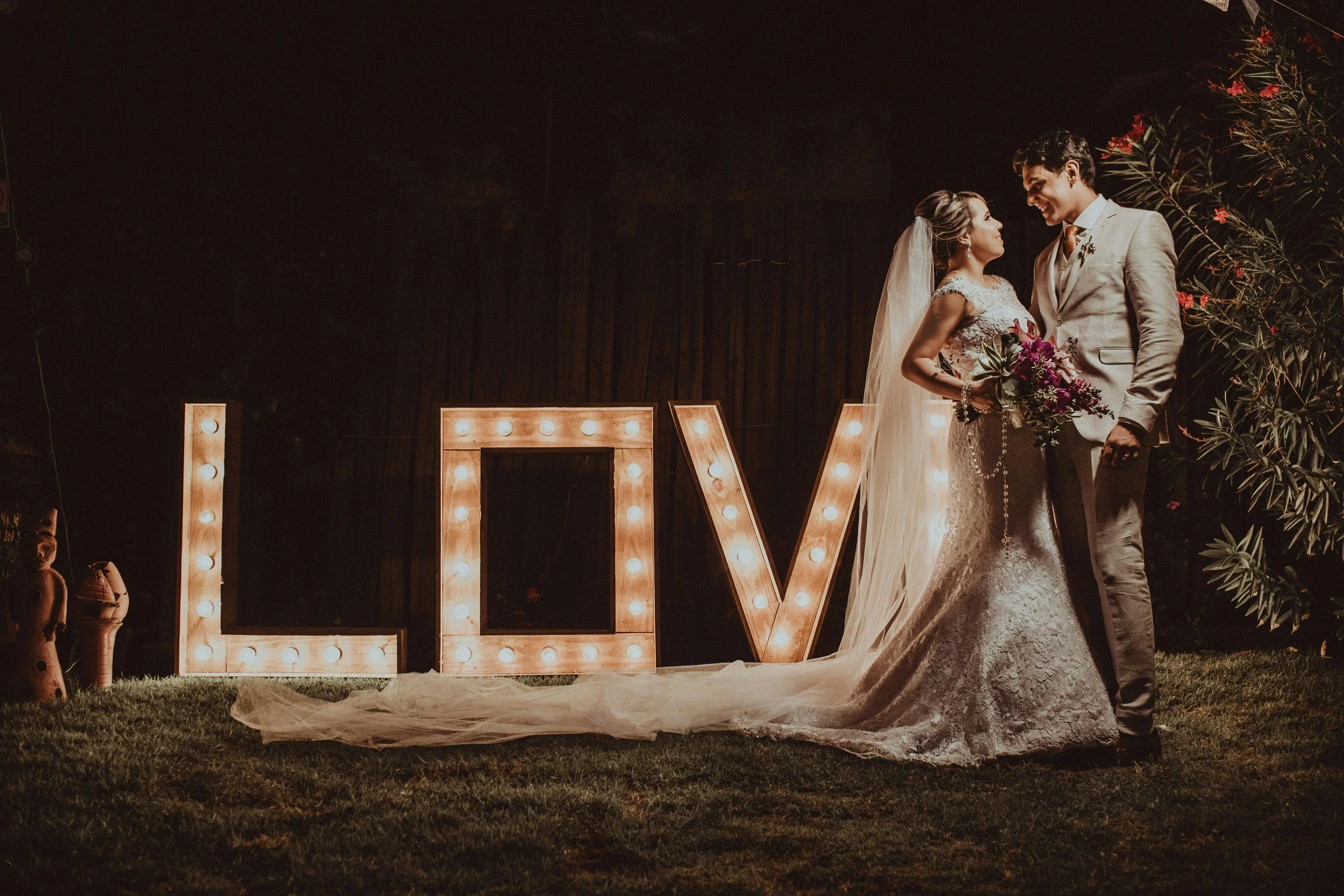 A bride and groom standing close together at night, smiling and looking at each other, near a large illuminated sign spelling "LOVE" with light bulbs, outdoors with trees and flowers in the background.