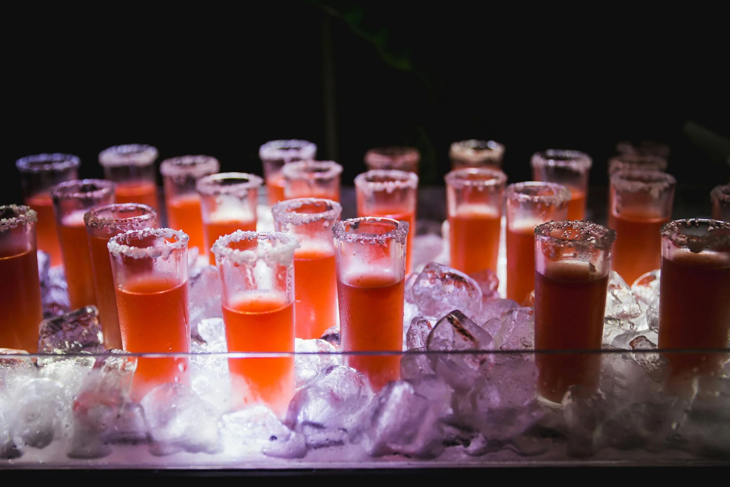 Multiple shot glasses filled with orange-colored drink, lined up on ice with salt on the rims, illuminated against a dark background.