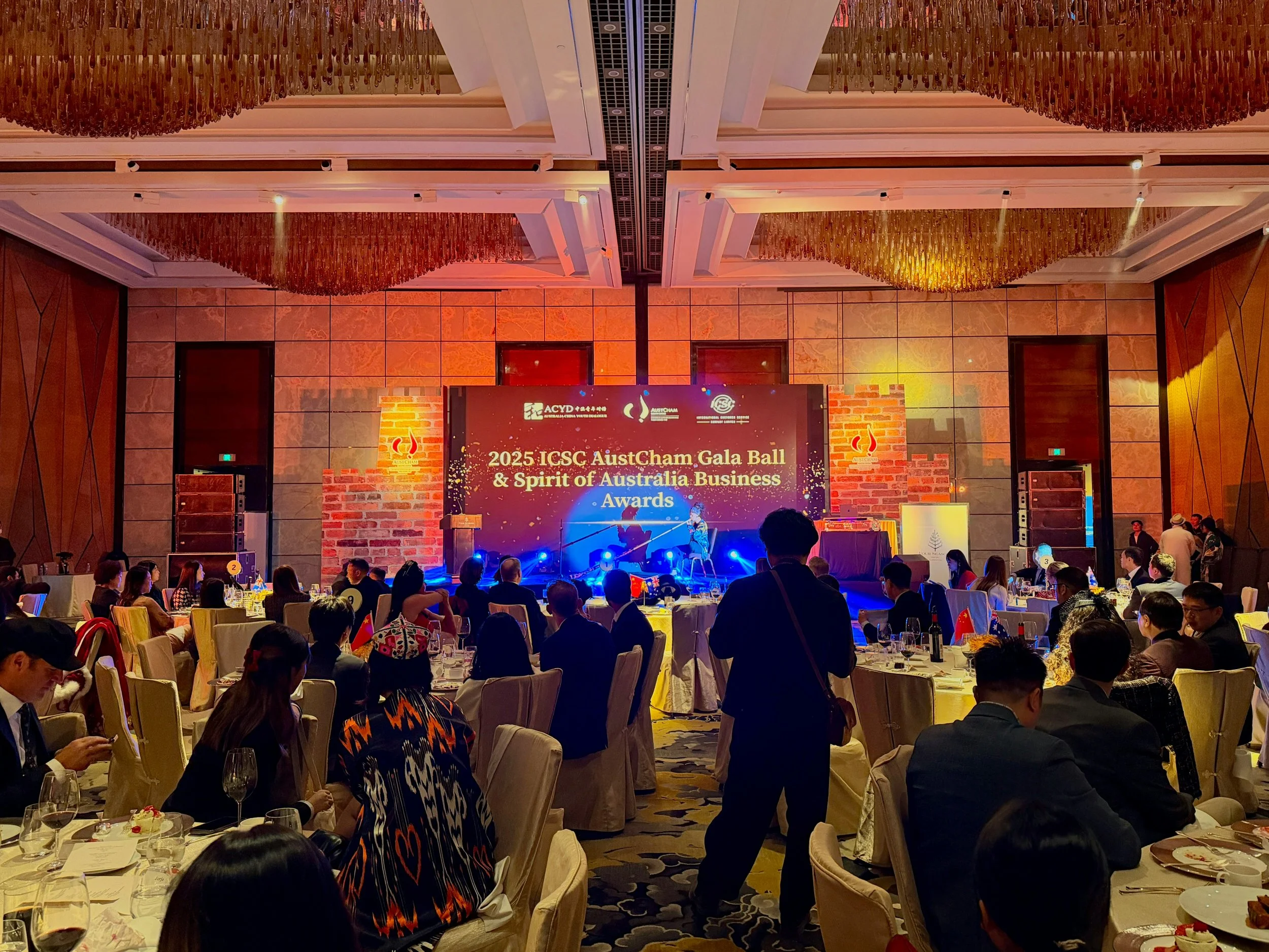 Attendees seated at round tables in a large banquet hall, watching a stage with a screen displaying event details for the 2025 ICSC AustCham Gala Ball & Spirit of Australia Business Awards, with speakers or performers on stage.