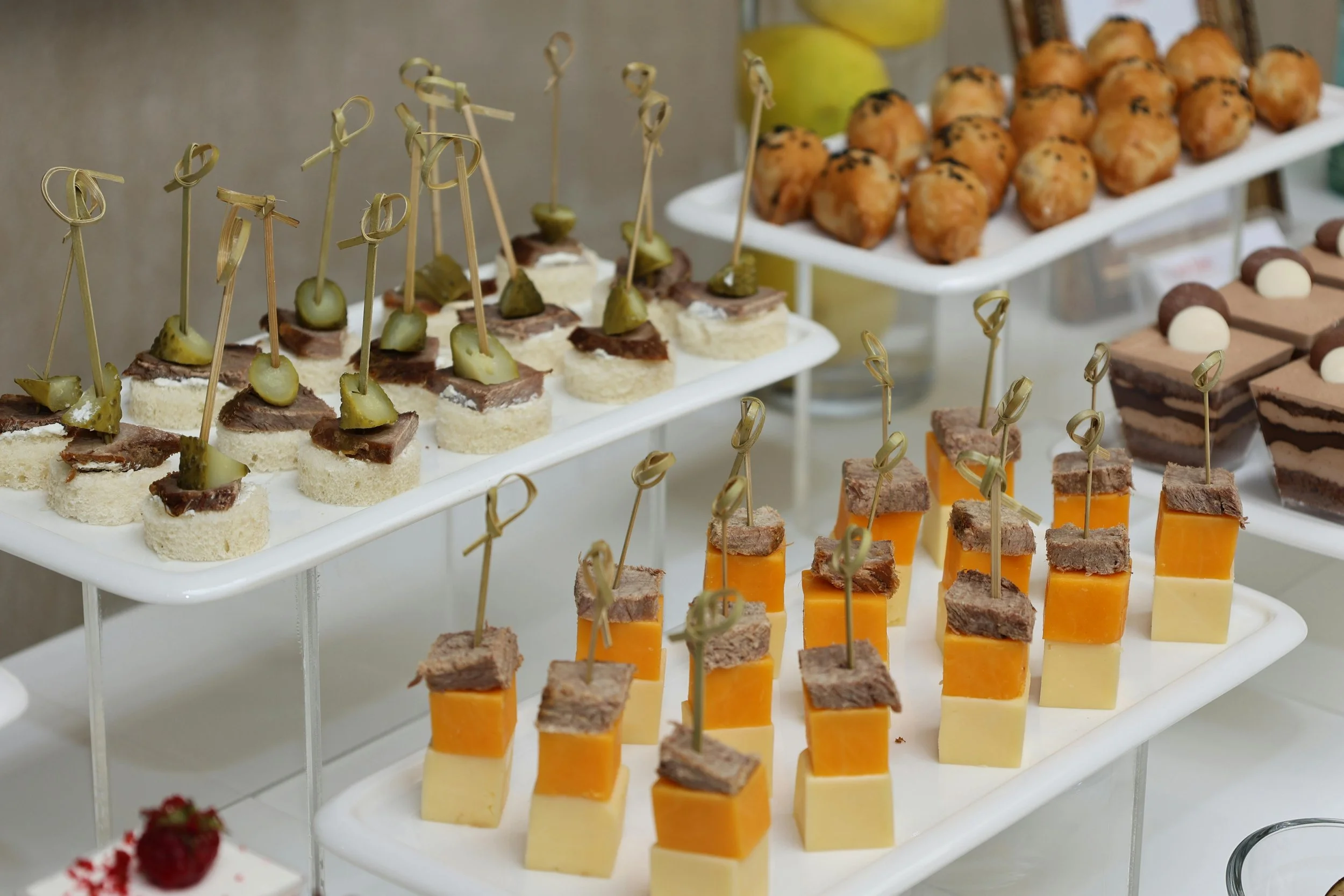 A variety of bite-sized appetizers arranged on tiered white serving trays, including cheese and meat cubes with toothpicks and small sandwiches with toppings.