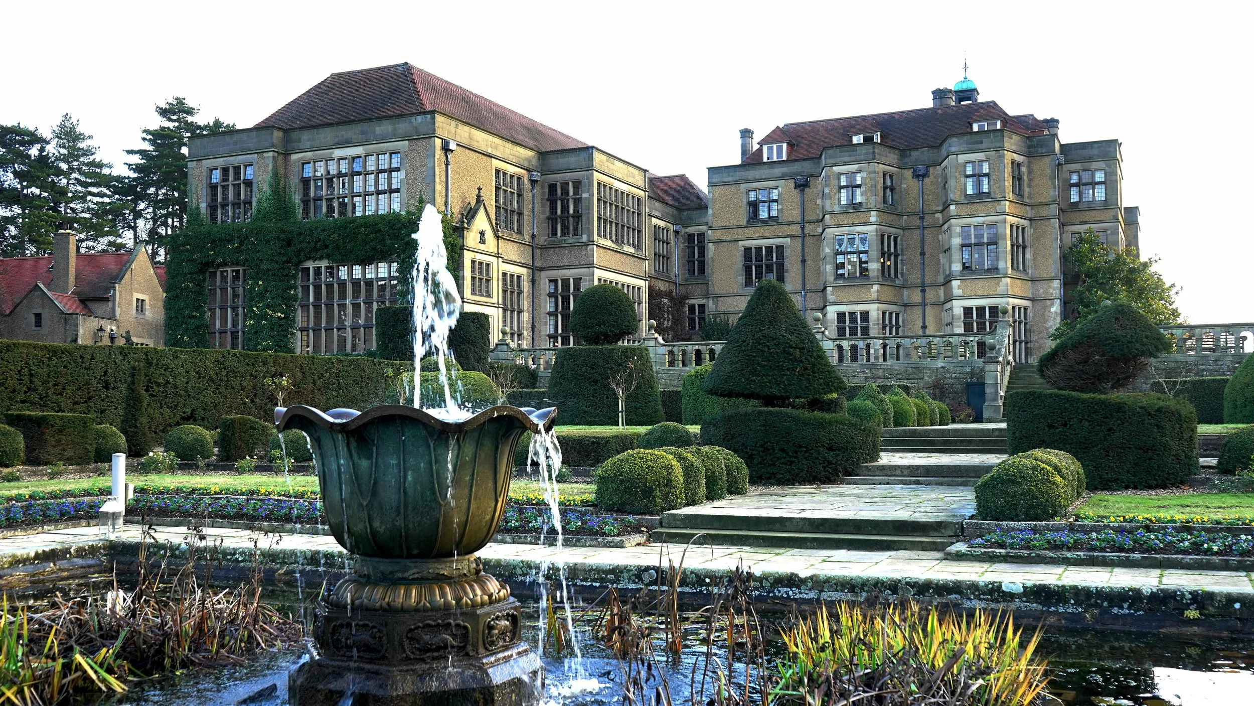 A historic stone mansion with large windows, surrounded by a well-manicured garden with trimmed bushes, a fountain, and stone steps leading up to the entrance.