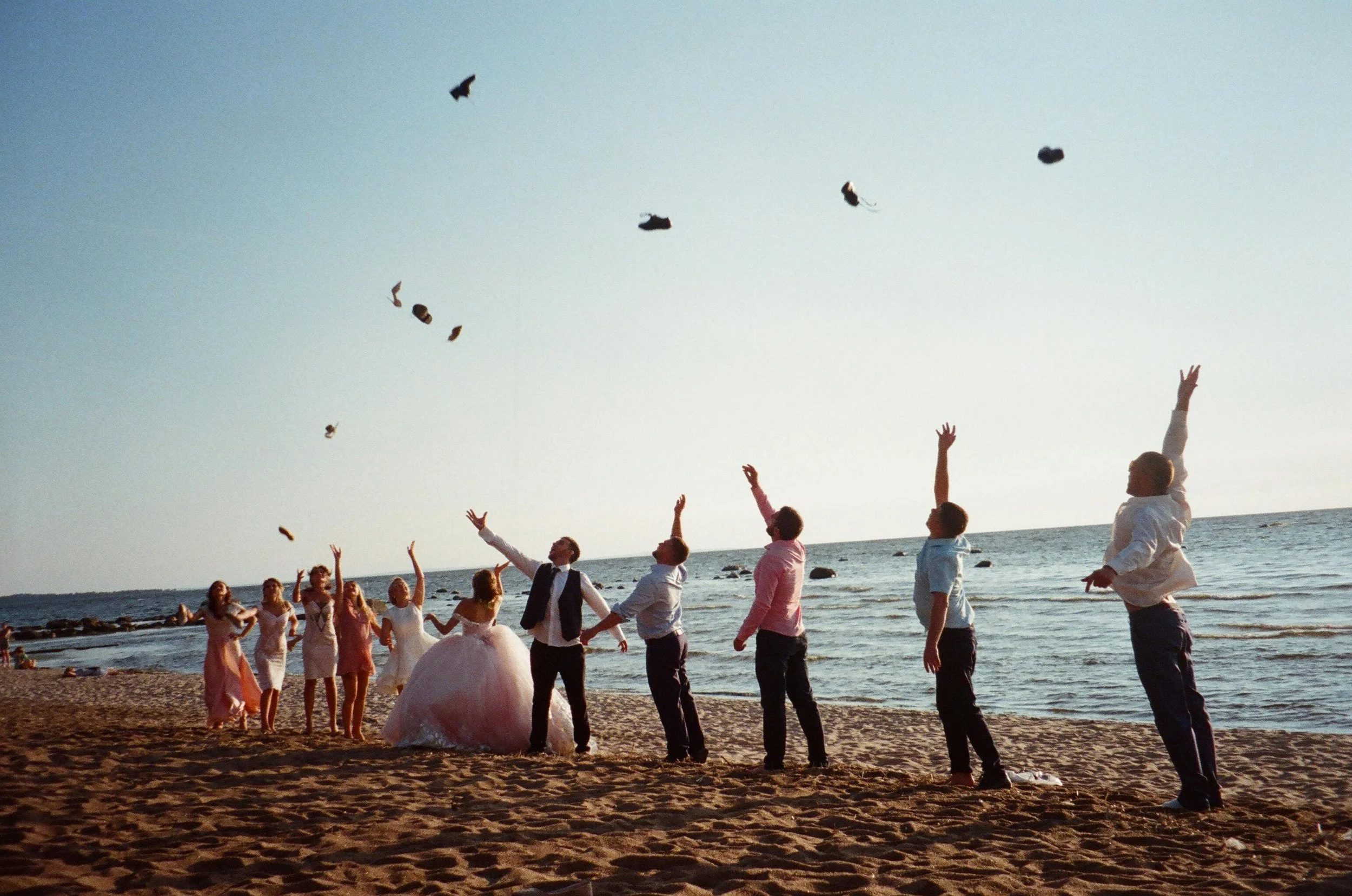 Group of people on the beach celebrating a wedding, throwing shoes into the air during sunset.