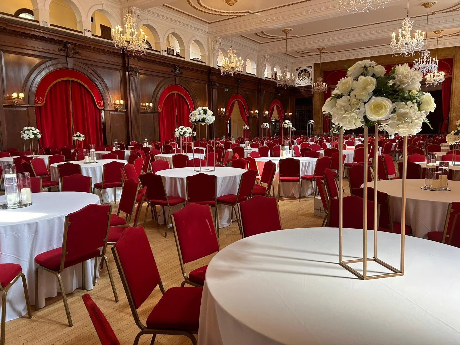 Elegantly decorated banquet hall with round tables covered in white cloths, red chairs, tall floral centerpieces with white roses, chandeliers, and red curtains.