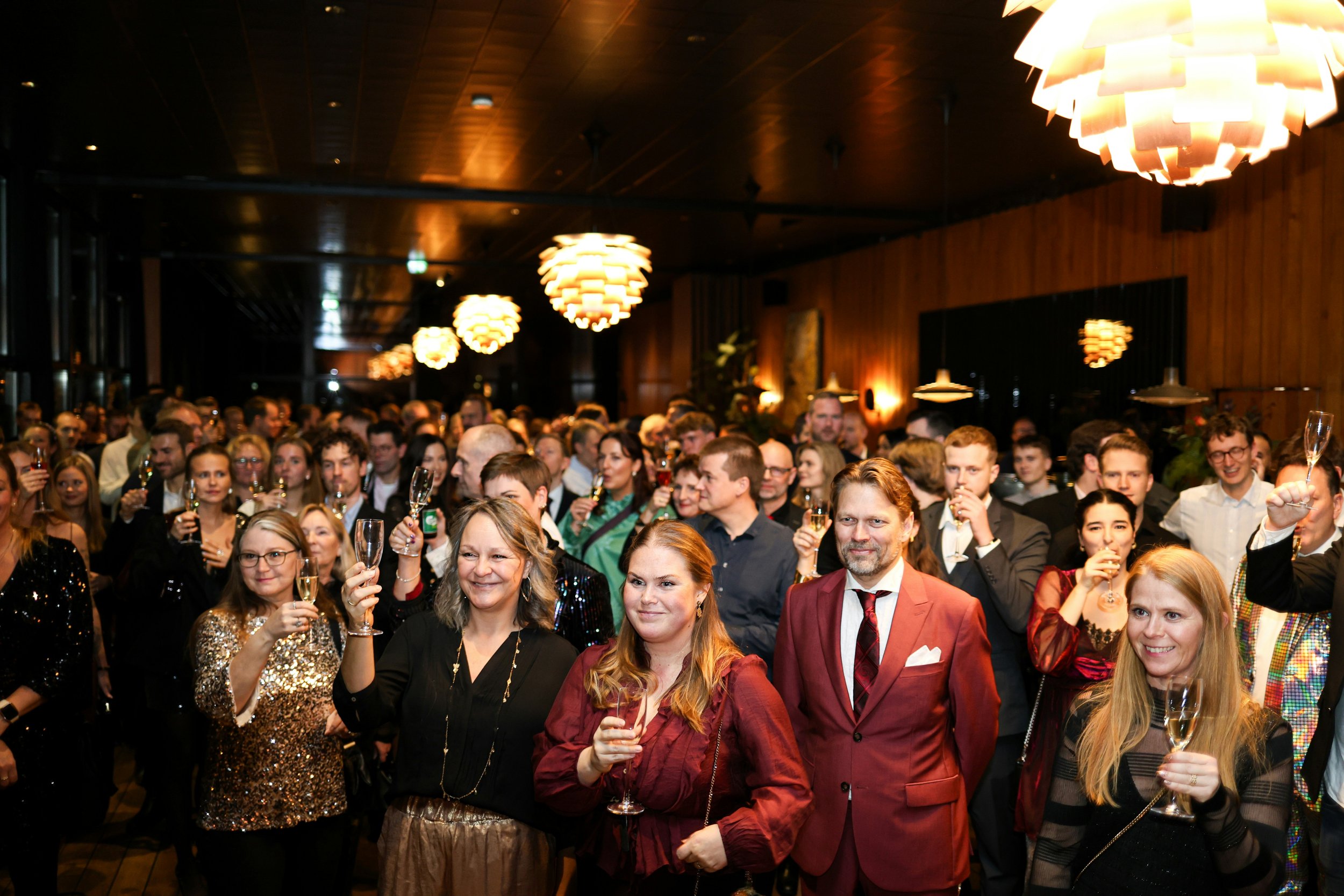 A crowd of people at an indoor celebration or party, many holding champagne glasses and raising them in a toast, in a warmly lit room with modern pendant lights and wooden walls.