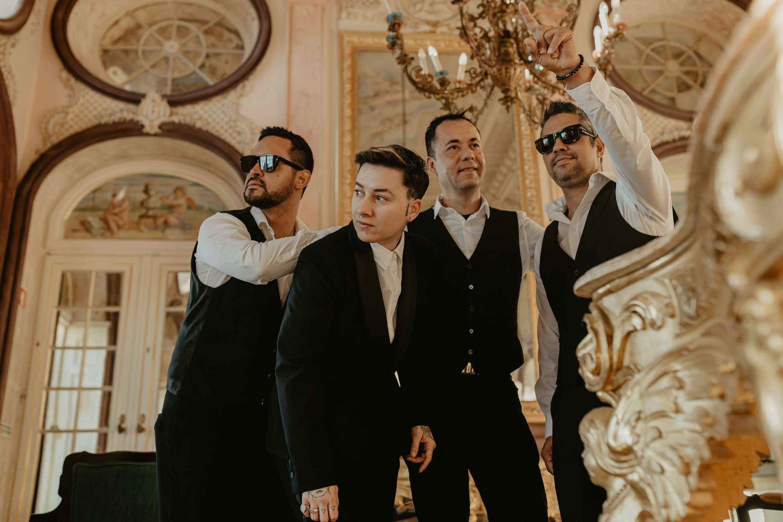 Four men dressed in formal black suits are standing in an ornate, gold-adorned room with chandeliers and decorative wall paintings. Two of them wear sunglasses, and one is making a gesture with his raised hand.