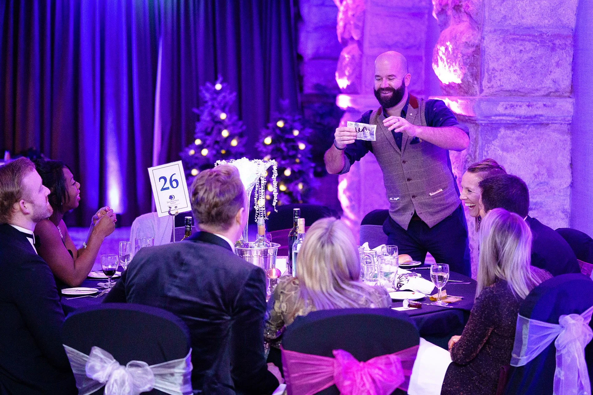 Man with a beard taking a picture with a phone at a formal dinner event with guests seated around a table decorated with Christmas trees and purple lighting.