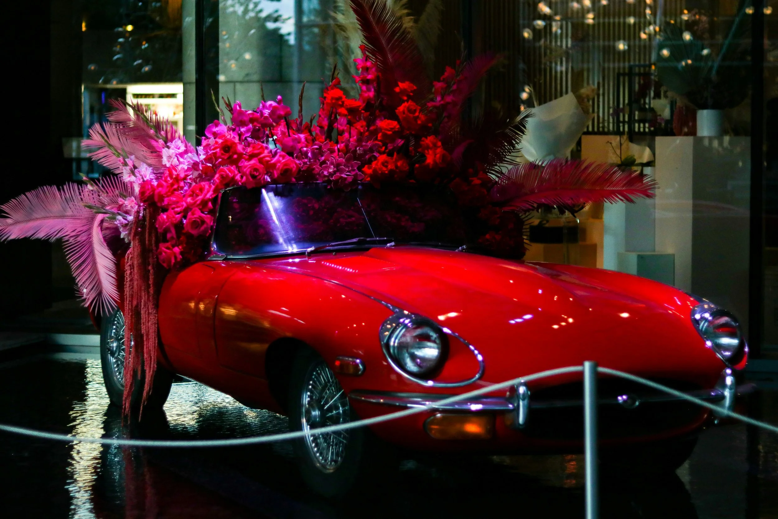 A vintage red sports car decorated with an elaborate arrangement of pink flowers and feathers, displayed behind a rope barrier in a modern indoor setting.