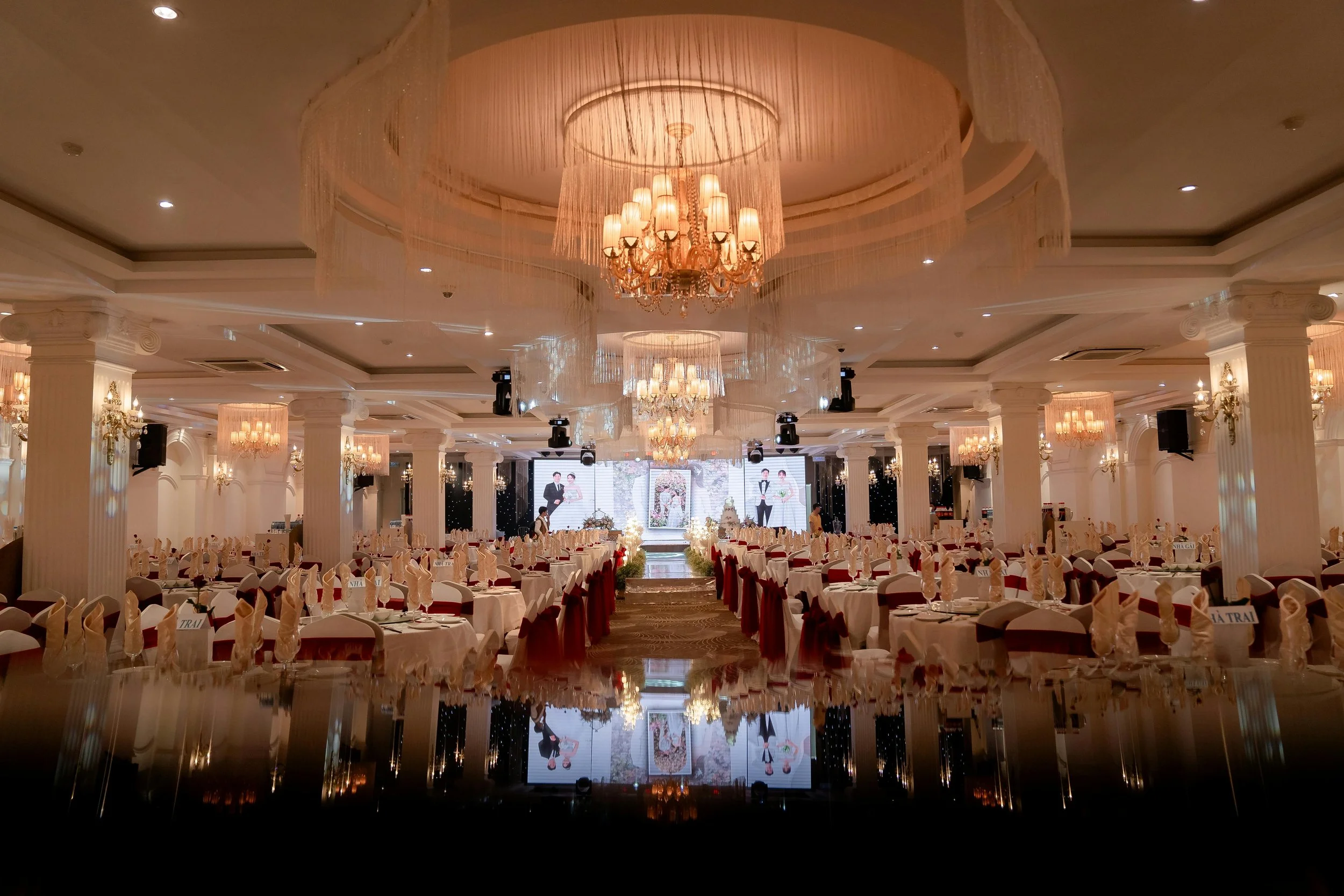 Elegant banquet hall decorated for a wedding reception with chandeliers, round tables with white tablecloths, and a stage with wedding photos.