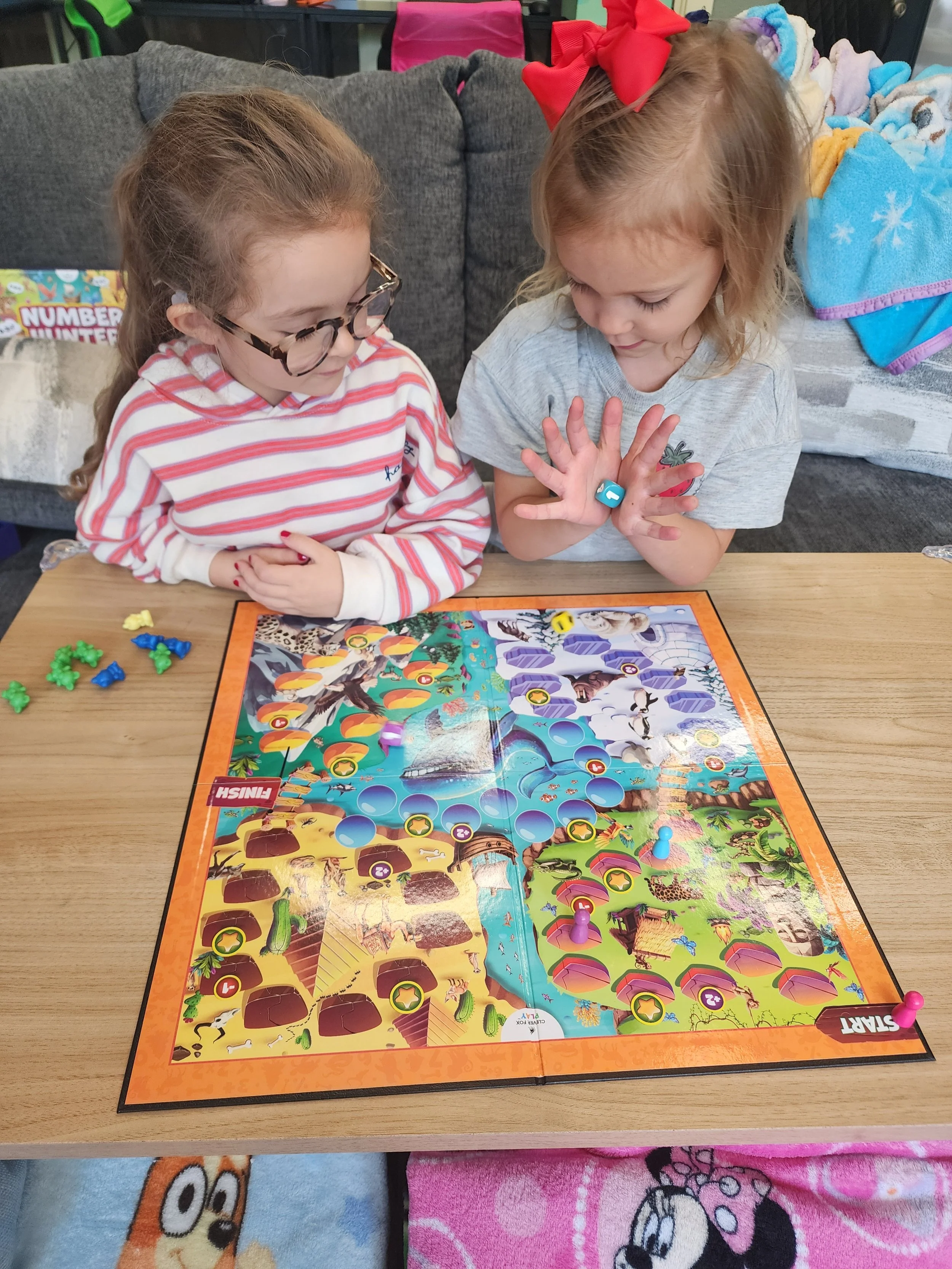 Two young girls playing a board game at a table. The game features colorful artwork and plastic game pieces. The girls are focused on the game, one wearing glasses and the other with a large red hair bow.