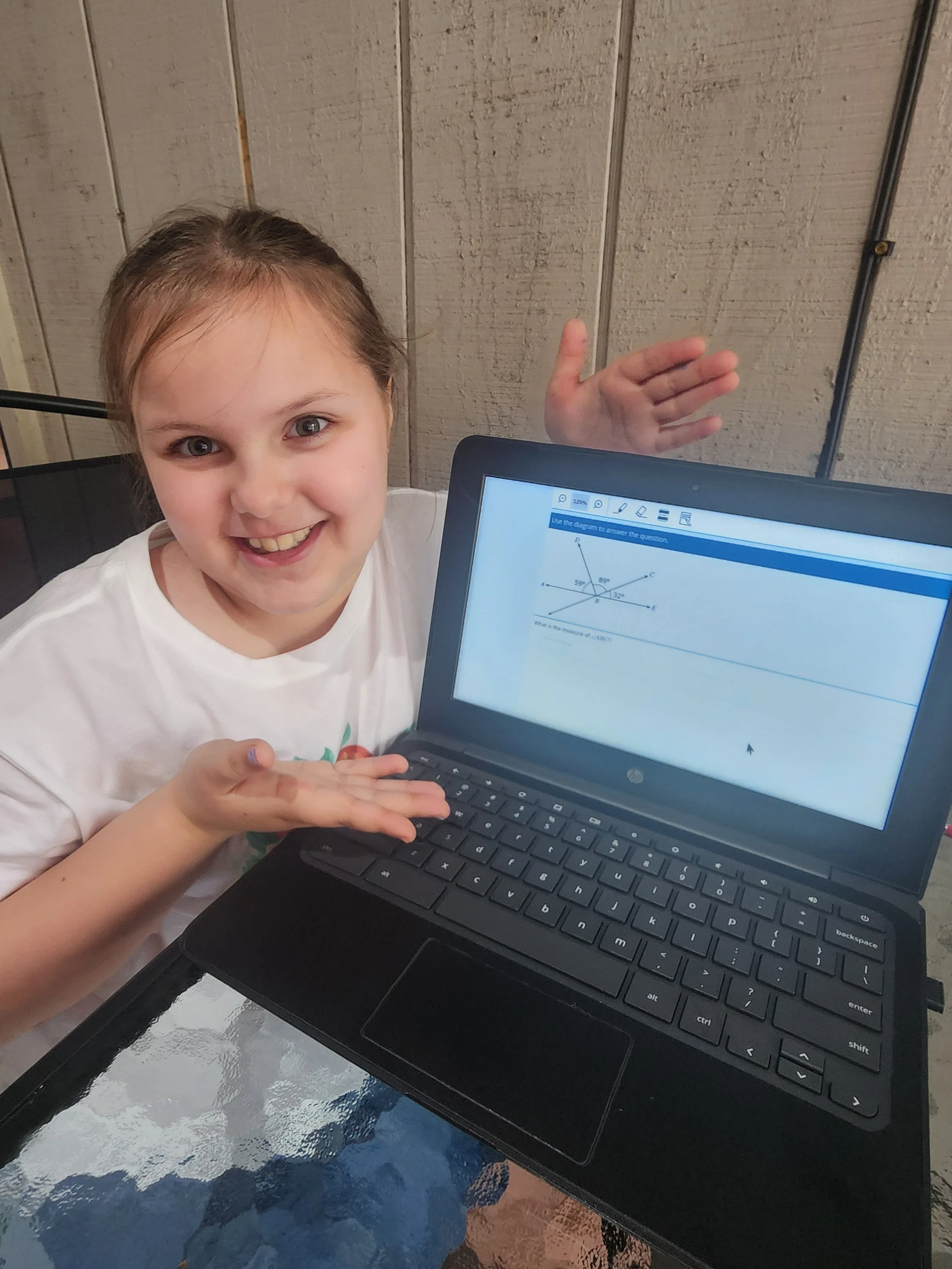 A young girl with brown hair smiling and in a white shirt, sitting at a table in front of a laptop displaying a math problem with a diagram of angles.
